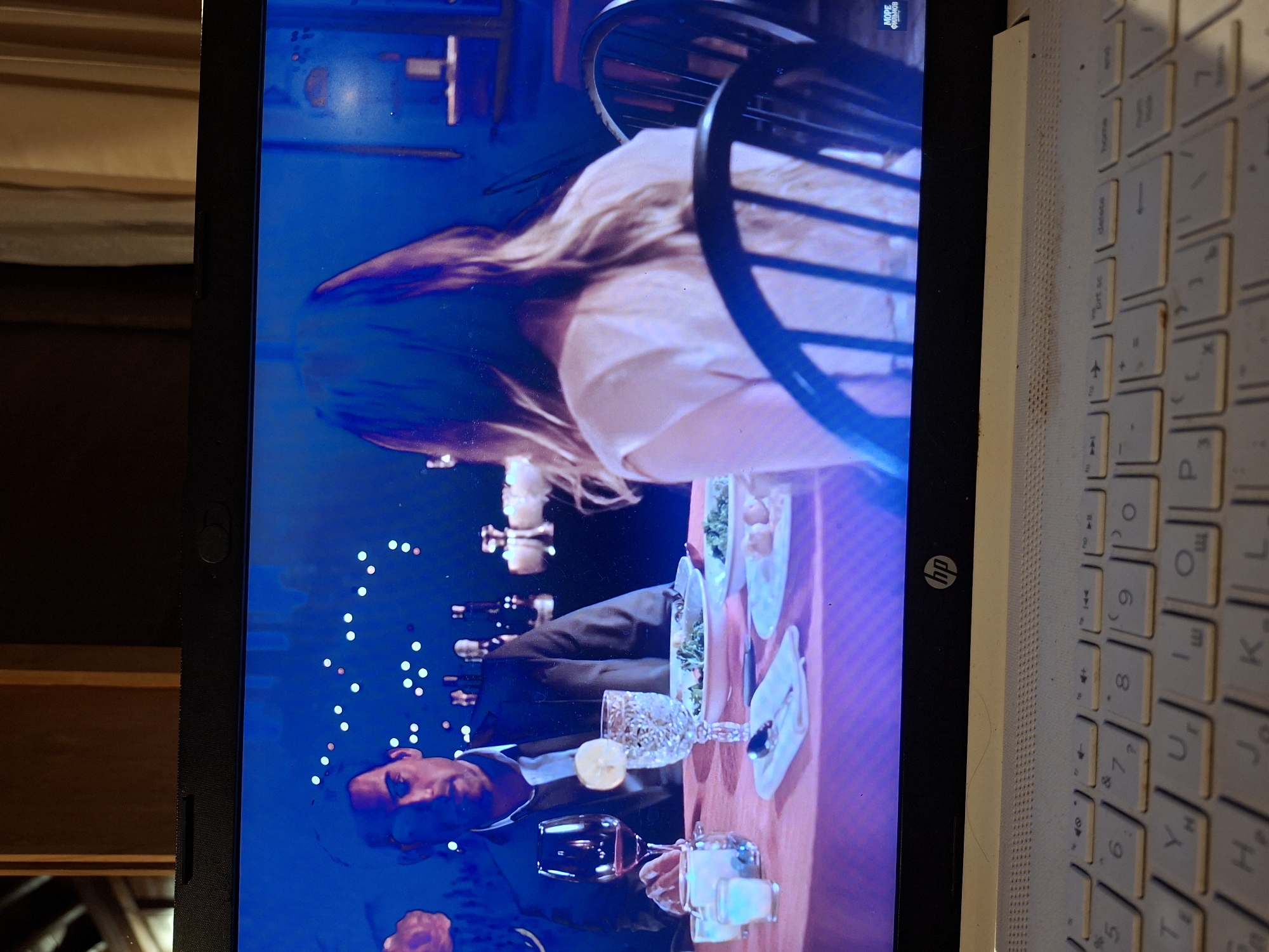 An image captures a close-up of a laptop displaying a scene from a video, situated in the Ubud District, Indonesia. The laptop screen shows an indoor dining setting, likely from a movie or TV show, with a blue ambient light. In the foreground of the screen, a man with short dark hair and a suit jacket is visible, holding a wine glass. A lemon slice garnishes a crystal-cut glass on the table in front of him, which is laden with various plates of food. To his right, a woman with long, light-colored hair is seated with her back partially towards the camera, also at the dining table. The table is set with multiple dishes, cutlery, and glasses, suggesting a formal meal.
The laptop itself is an HP brand, with its logo visible on the bottom bezel of the screen. The open laptop is positioned next to a light-colored keyboard that features both Latin (QWERTY layout) and Cyrillic characters, indicating a non-English language setting. Keys like "home," "delete," and "ctrl sc" are discernible. The keyboard surface appears to have some dust or debris. In the upper left corner of the laptop screen, the text "MOPE ФИЛЬМОВ" (Russian for "SEA OF FILMS" or "MANY FILMS") is visible, indicating the video source. To the left of the laptop, a dark, textured object, possibly a piece of furniture, is partially visible. The overall ambiance of the video scene appears to be evening or night due to the lighting.