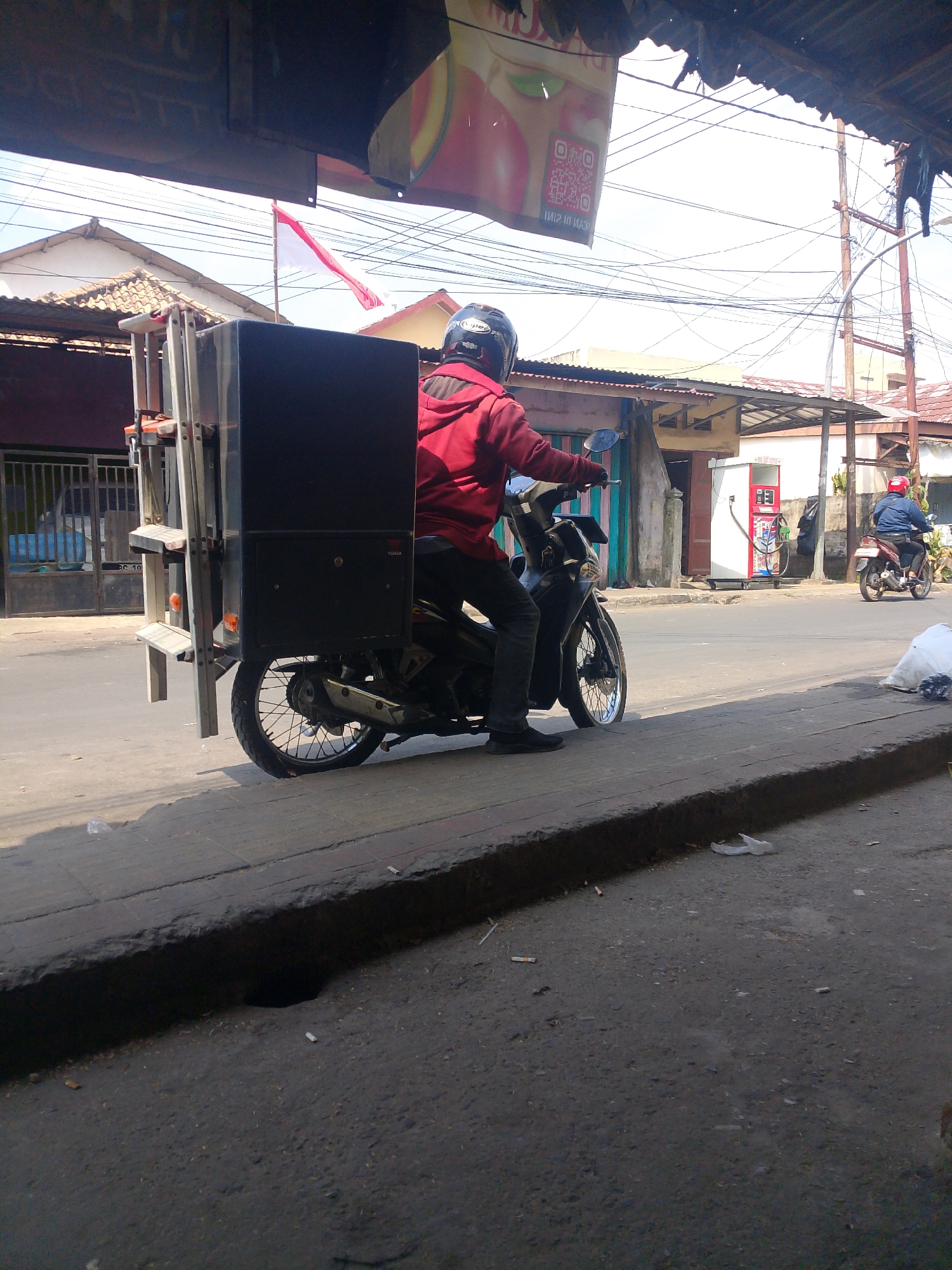 A side view of a person wearing a red jacket and a helmet riding a motorcycle with a large black box attached to the back, moving along a street in Palembang, Indonesia. The motorcycle has a license plate that is partially visible. In the background, there are buildings with tiled roofs, power lines, and a red and white flag. Another person on a motorcycle is seen further down the street near a gas station. The foreground shows a paved sidewalk and a road. The weather appears to be sunny.
