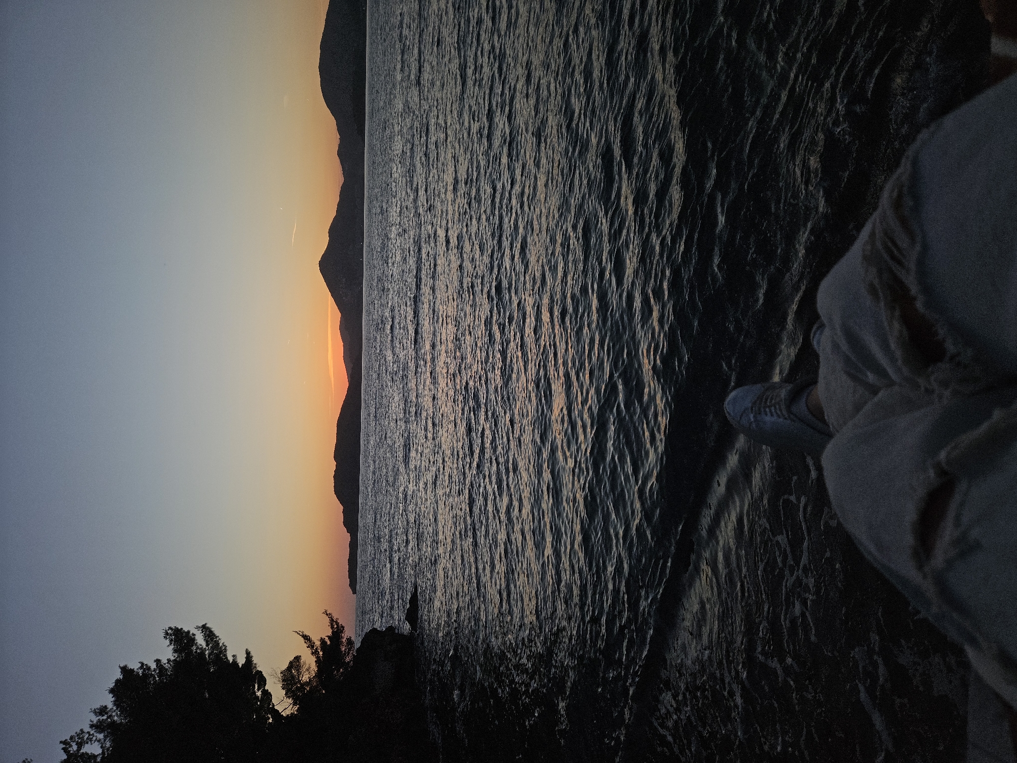 The image captures a serene coastal landscape during either sunset or sunrise. A person's lower leg and foot, wearing a light-colored shoe and ripped jeans, are visible on the bottom right, suggesting they are seated or standing near the water's edge, observing the scene.
The horizon line extends across the middle of the image, marked by a range of dark, silhouetted mountains or landmasses. Above the mountains, the sky displays a beautiful gradient, transitioning from a deep blue-grey at the top to vibrant orange and yellow hues near the horizon, where the sun is either setting or rising.
A large body of water, likely the sea, occupies the midground, its surface covered in subtle ripples that catch and reflect the ambient light. On the bottom left, dark, silhouetted trees or bushes are visible, framing the foreground.
The overall atmosphere is peaceful and contemplative, enhanced by the warm, soft light of the sun and the calm appearance of the water. The scene depicts a tranquil moment in a natural setting. This view is of Sveti Stefan, Montenegro. There is no visible text in the image.