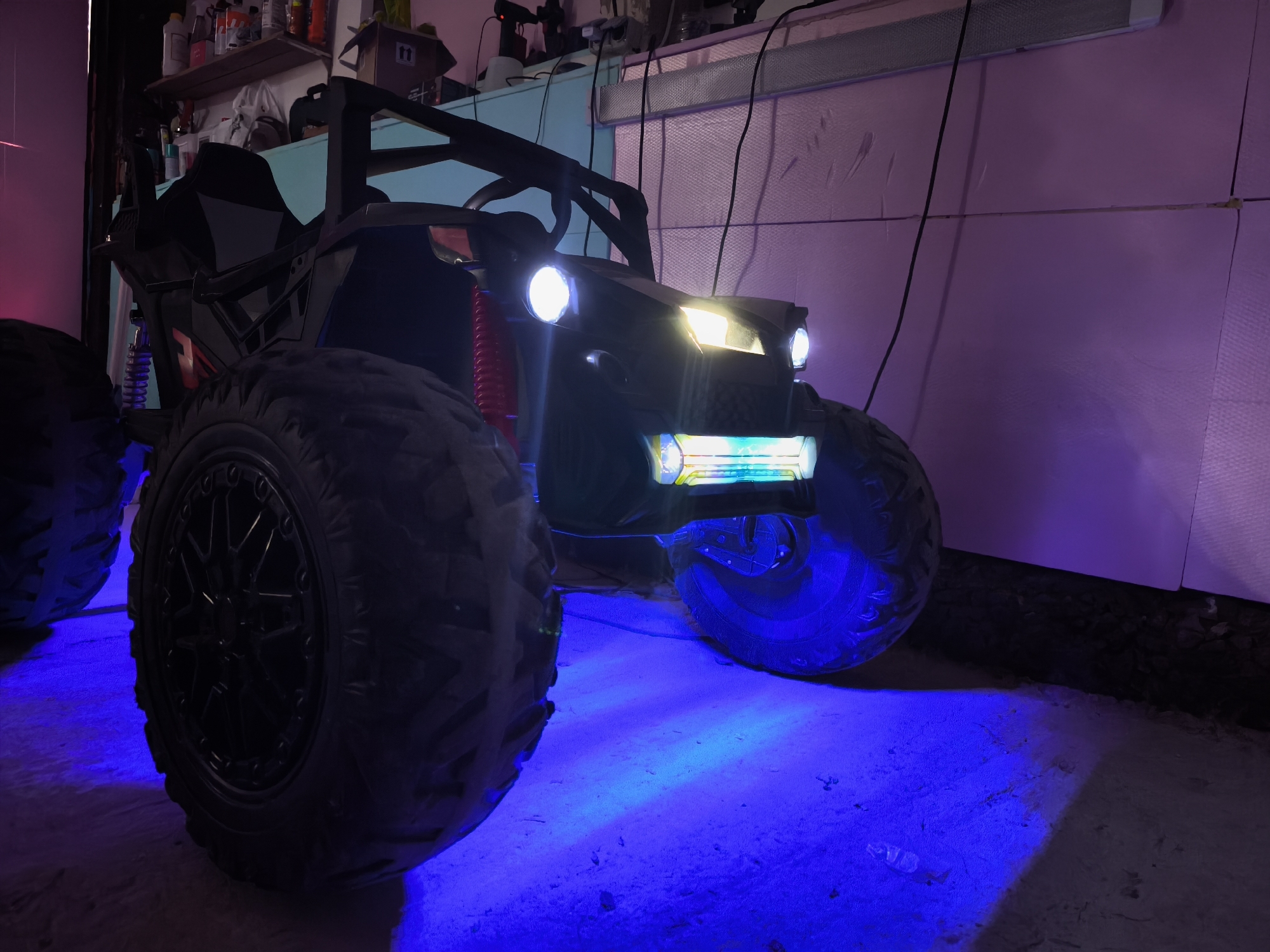 An indoor, low-angle shot features a large, dark-colored off-road vehicle, possibly a ride-on toy or a substantial RC car, positioned centrally. The vehicle has prominent, knobby tires with black multi-spoke rims. Its headlights emit bright white and yellow light, while a lower light bar glows with blue and green hues. These lights cast a vivid blue illumination onto the concrete or dirt floor directly in front and underneath the vehicle.
In the background, shelves are visible, holding various items such as bottles and boxes, suggesting a garage, workshop, or storage area. A light pinkish-purple hue illuminates the wall behind the vehicle, which appears to be constructed from textured panels, possibly insulation boards or foamboard. Several black cables hang down in the upper background.
The scene is dominated by artificial and colored lighting, creating a dramatic and moody atmosphere. The vehicle is stationary, with its lights actively on. A faint "11" is visible on a brown cardboard box on the top shelf in the background. The low perspective emphasizes the vehicle's size and its illuminated presence.