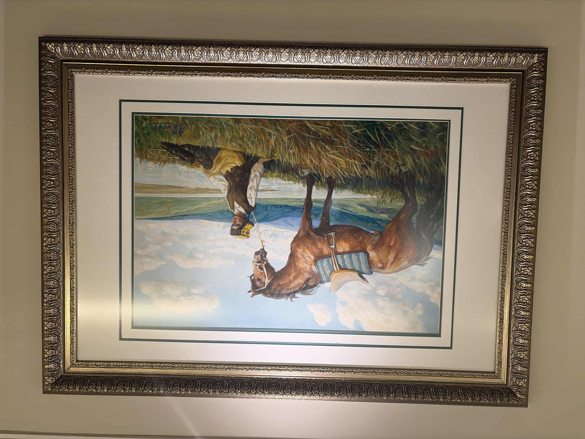 A framed oil painting depicts a brown horse standing in a grassy field, with a young woman in traditional clothing sitting beside it. The woman is wearing a brown vest over a white shirt and a yellow skirt, with a decorative yellow and brown hat. The horse is saddled with a blue and white patterned blanket. Behind them, a body of water stretches towards a rolling landscape of green hills and mountains under a blue sky with white clouds. The painting is signed in the bottom right corner. The frame is ornate and gold-colored. The painting is displayed on a plain, light-colored wall.