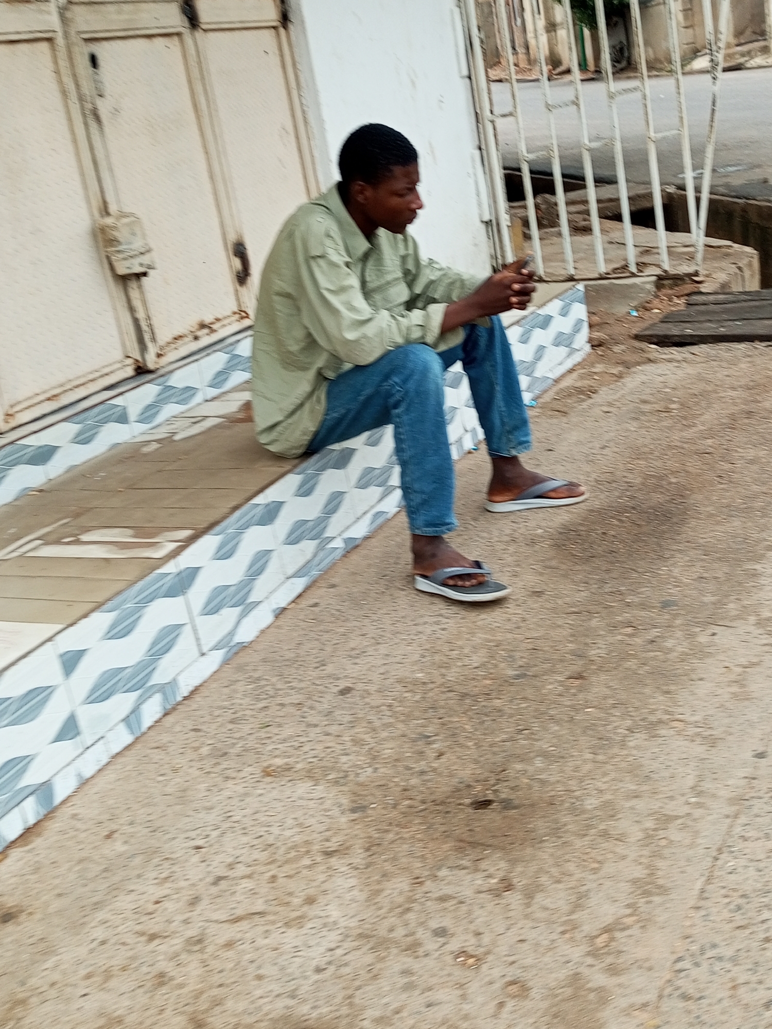 A young Black man wearing a light green collared shirt and blue jeans sits on a tiled ramp outside a building. He is wearing flip-flops. His hands are clasped together in his lap. Behind him is a large, weathered metal door. To the right of the door is a white wall and a metal fence that separates the walkway from a street. The ground in front of the ramp is concrete.
