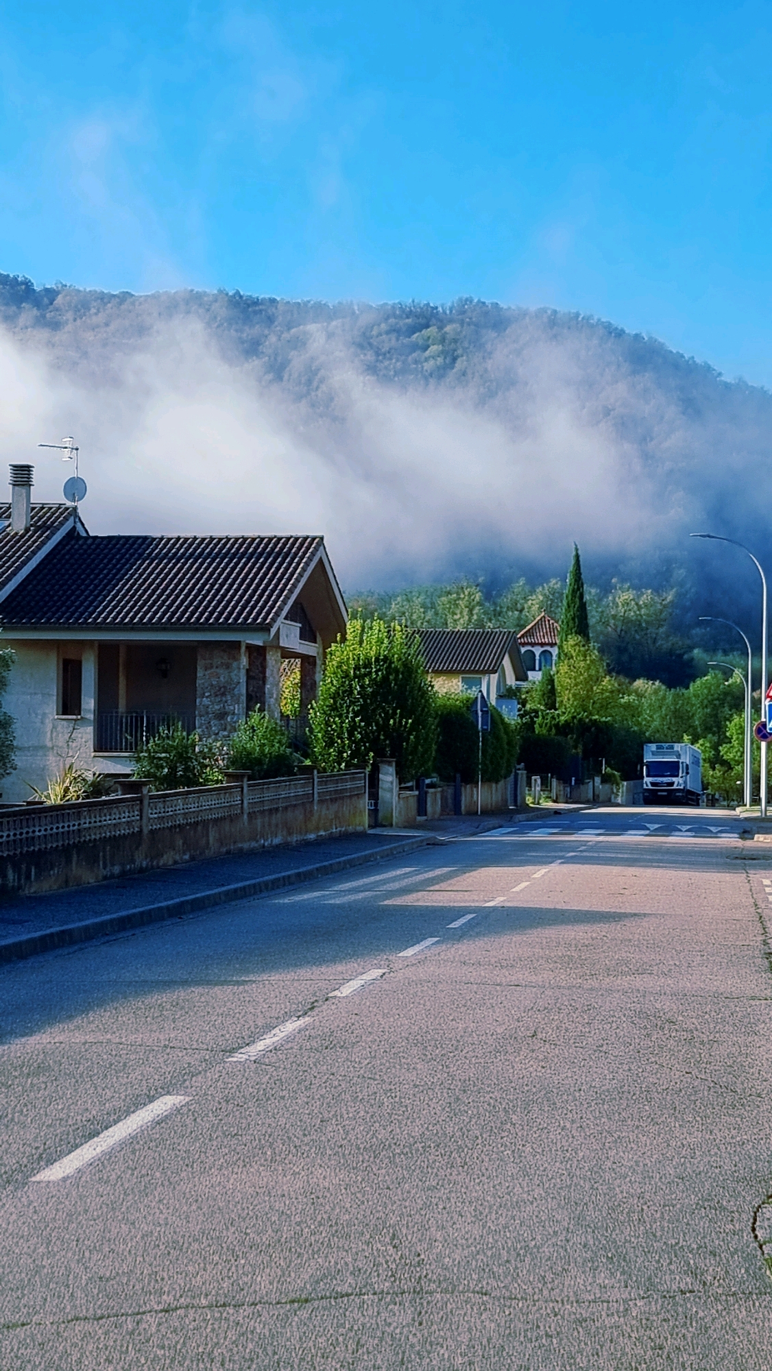 The image depicts a street scene in Sant Joan Les Fonts, Spain, characterized by a bright, clear blue sky. A gently winding asphalt road occupies the foreground and extends into the midground, marked with dashed white lines. On the left side of the road, a low, decorative stone wall borders a residential property. Behind the wall are houses with dark tile roofs. One house features a light-colored facade with stone accents and a covered porch. Mature green trees and shrubs are visible in front of and between the houses. A tall, slender evergreen tree stands out amongst other greenery further down the street.
In the background, a densely wooded mountainside rises, partially obscured by wisps of white mist that hug its lower slopes. The mist creates a soft, ethereal effect, contrasting with the sharp details of the foreground. A white box truck is parked on the right side of the road in the midground. Streetlights are visible along the right side of the road, and a traffic sign is also present. The overall impression is of a quiet, residential area on a clear, slightly misty morning.