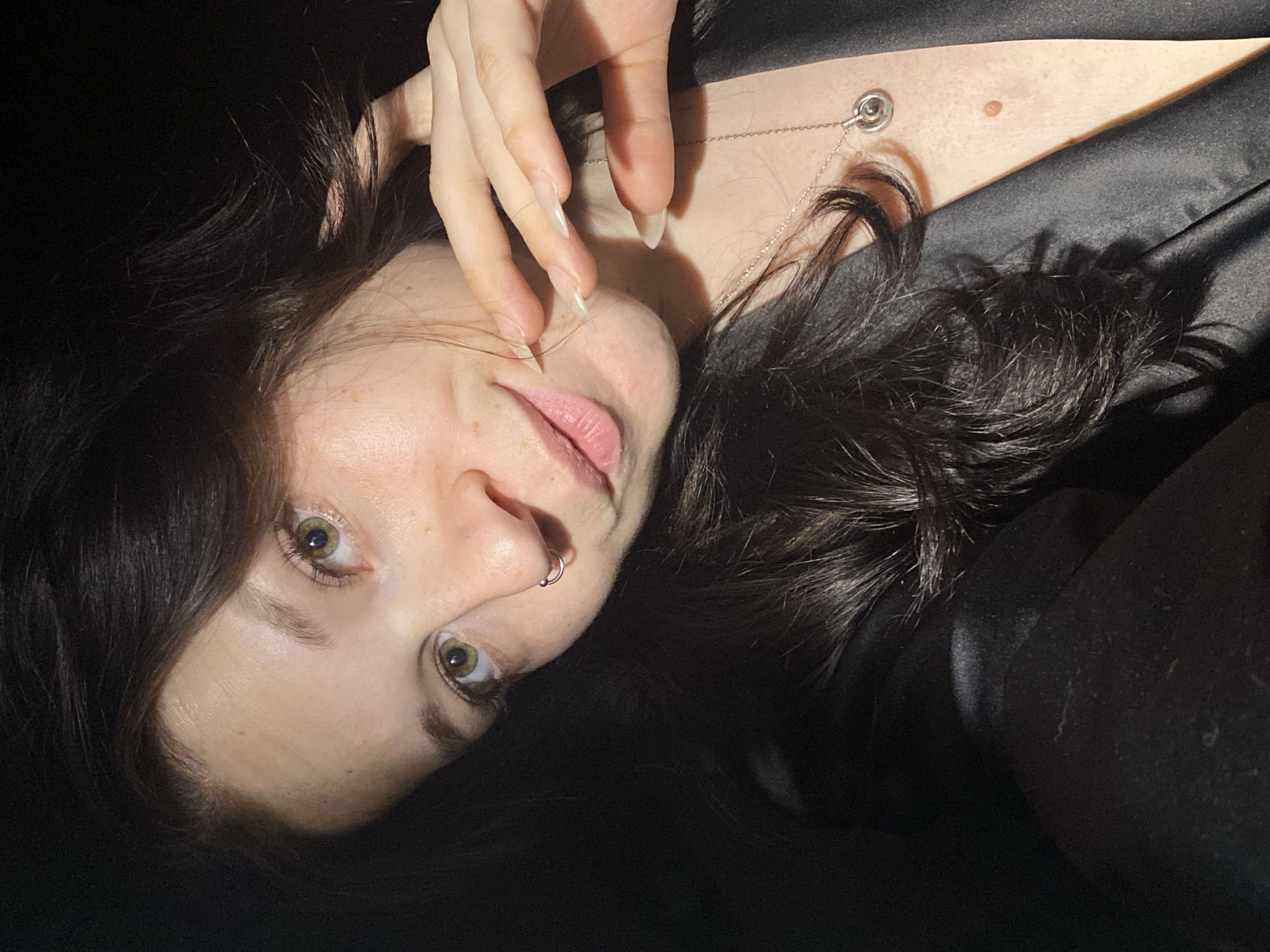 A close-up, rotated image depicts a fair-skinned individual with dark hair. The person has light eyes, a nose ring, and pale pink lips. Their hand is near their face. A necklace with a round pendant is visible on the person's neck/chest. The background is mostly dark, with a hint of what seems to be dark fabric. The location is not identifiable from this image.