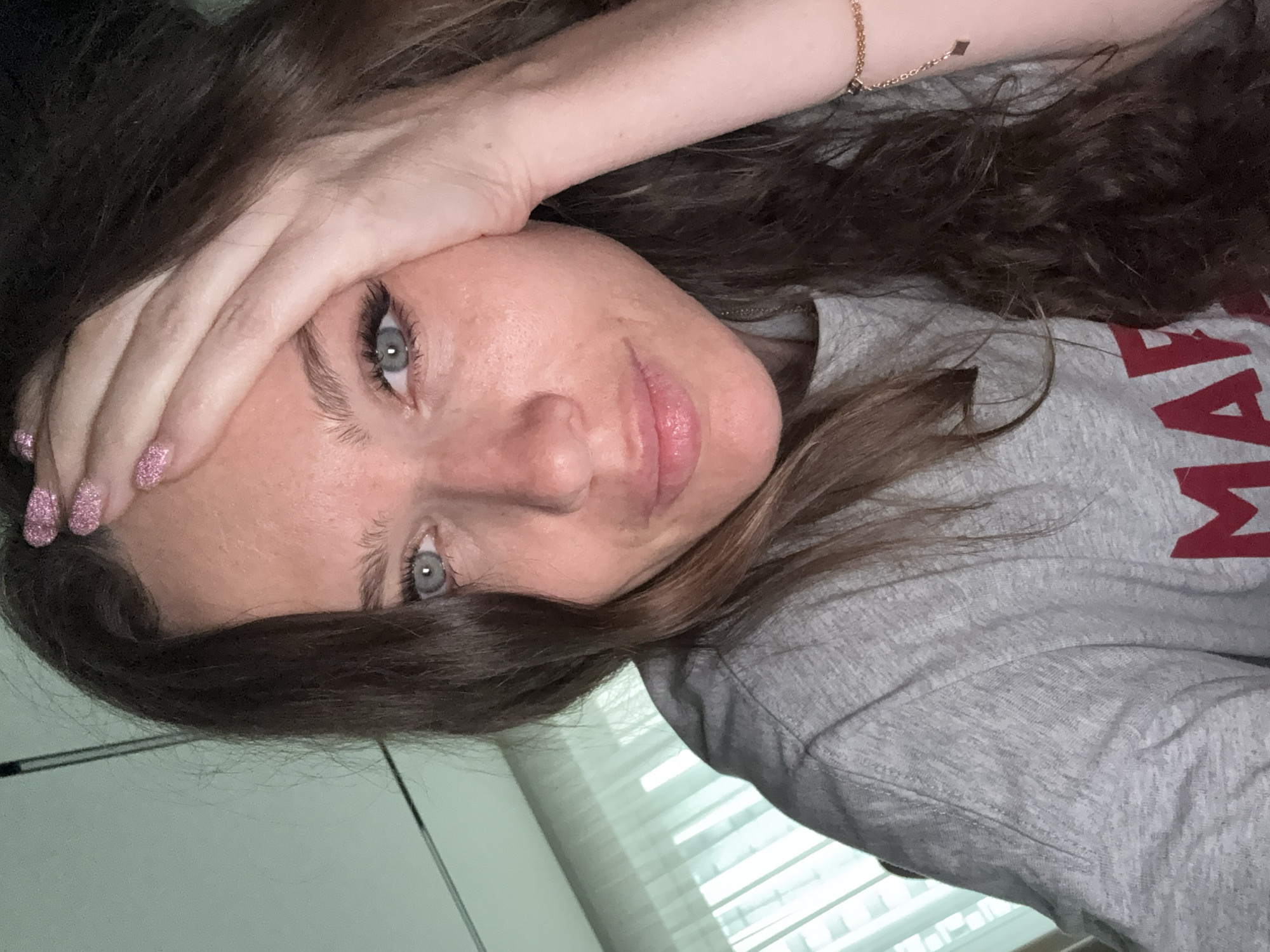 A woman with long, dark hair and light blue eyes is lying down, looking at the camera. Her right hand is on her forehead. She is wearing a grey sweatshirt with red lettering. A gold bracelet is visible on her right wrist. The background includes a window with blinds and a light-colored wall. The location is not discernible as Khimki, Russia, based on the visible elements.