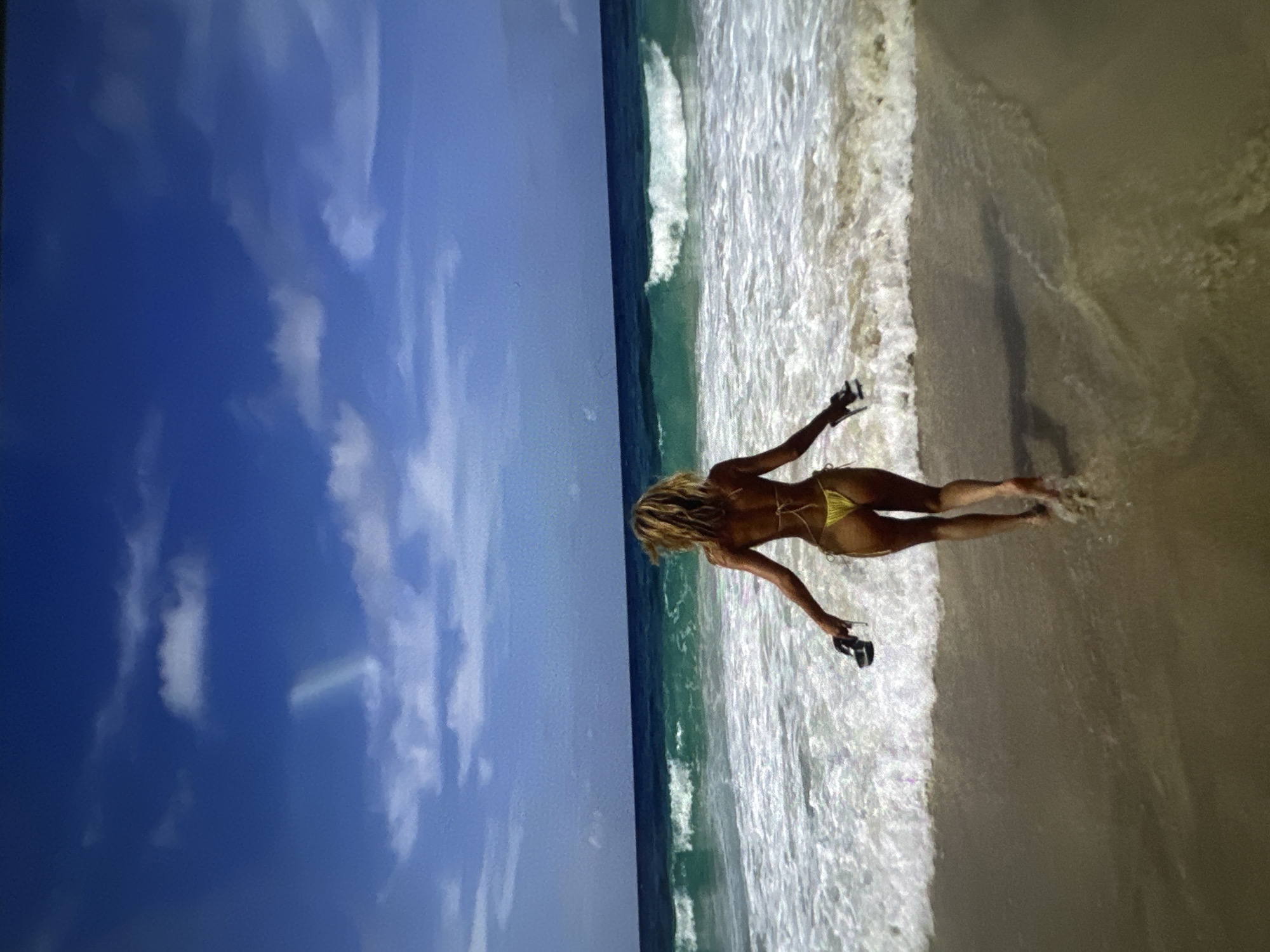 A photograph shows a woman in a yellow bikini walking on a beach. The scene includes a blue sky with clouds, the ocean, and the sandy shore. The woman is holding what appears to be sandals in her left hand. The exact location and city are unknown.