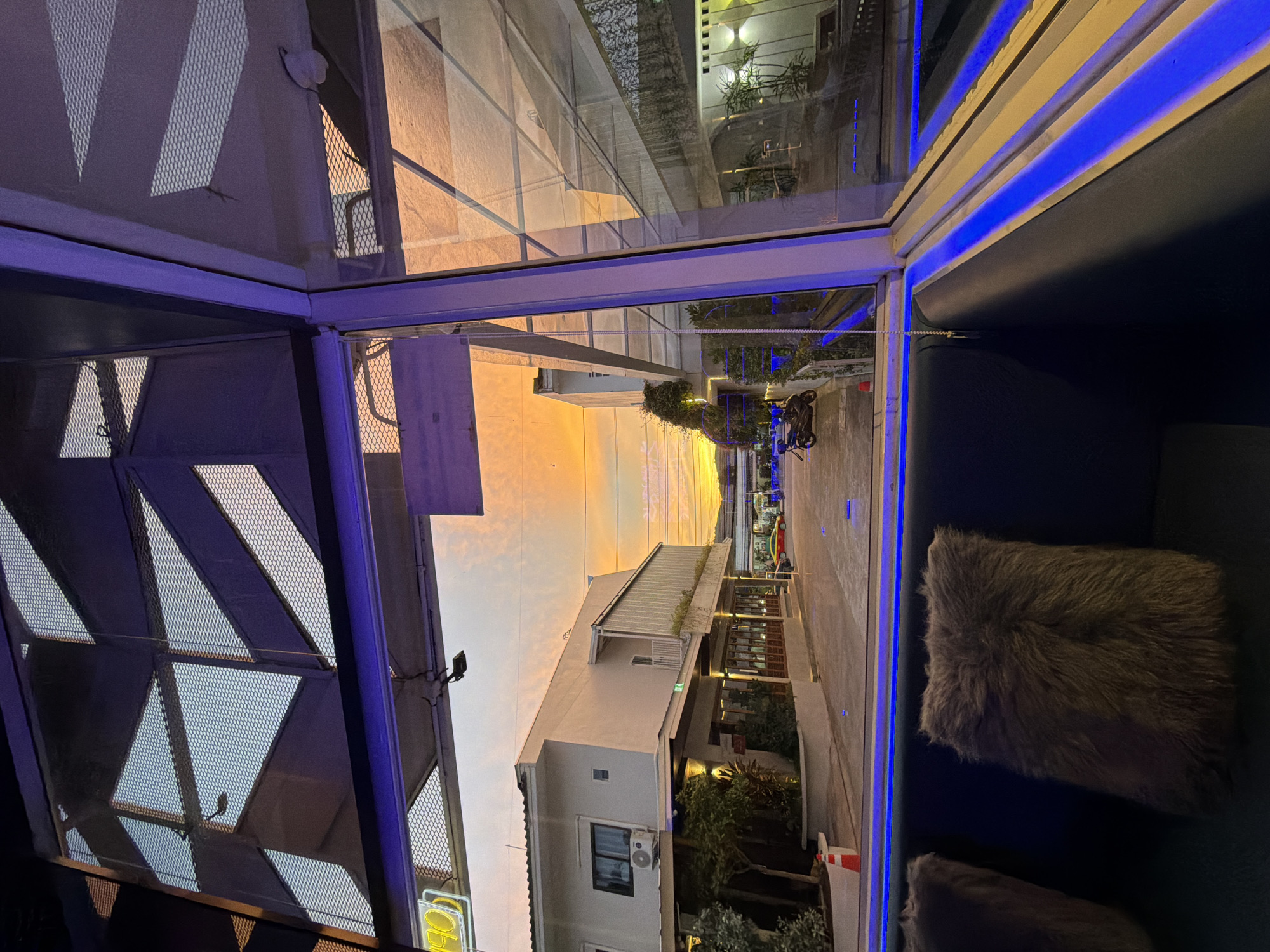 The image, taken at dusk in an urban setting, showcases a view from a dimly lit interior looking out through a large glass window. The interior features dark seating with plush, gray fur pillows and is illuminated by vibrant blue LED strip lighting along the window frame.
The exterior scene depicts a street with buildings on either side. On the left, a modern, multi-story building with white and beige facades is visible. It has several windows, one with an air conditioning unit, and a balcony. The ground floor appears to be a commercial space with large windows and wooden accents. A white traffic cone stands on the pavement in front of this building. Across the street, another building with similar modern architecture is visible, featuring rows of potted plants and white lights. A motorcycle is parked on the street. In the distance, a red car is driving away.
The sky is dramatic, with a vibrant orange and yellow sunset casting a warm glow. The sky is partially obscured by overhead structures with a geometric, lattice-like design, which create interesting patterns of light and shadow. The overall impression is a contemporary urban environment at twilight, viewed from a comfortable, well-lit indoor space.