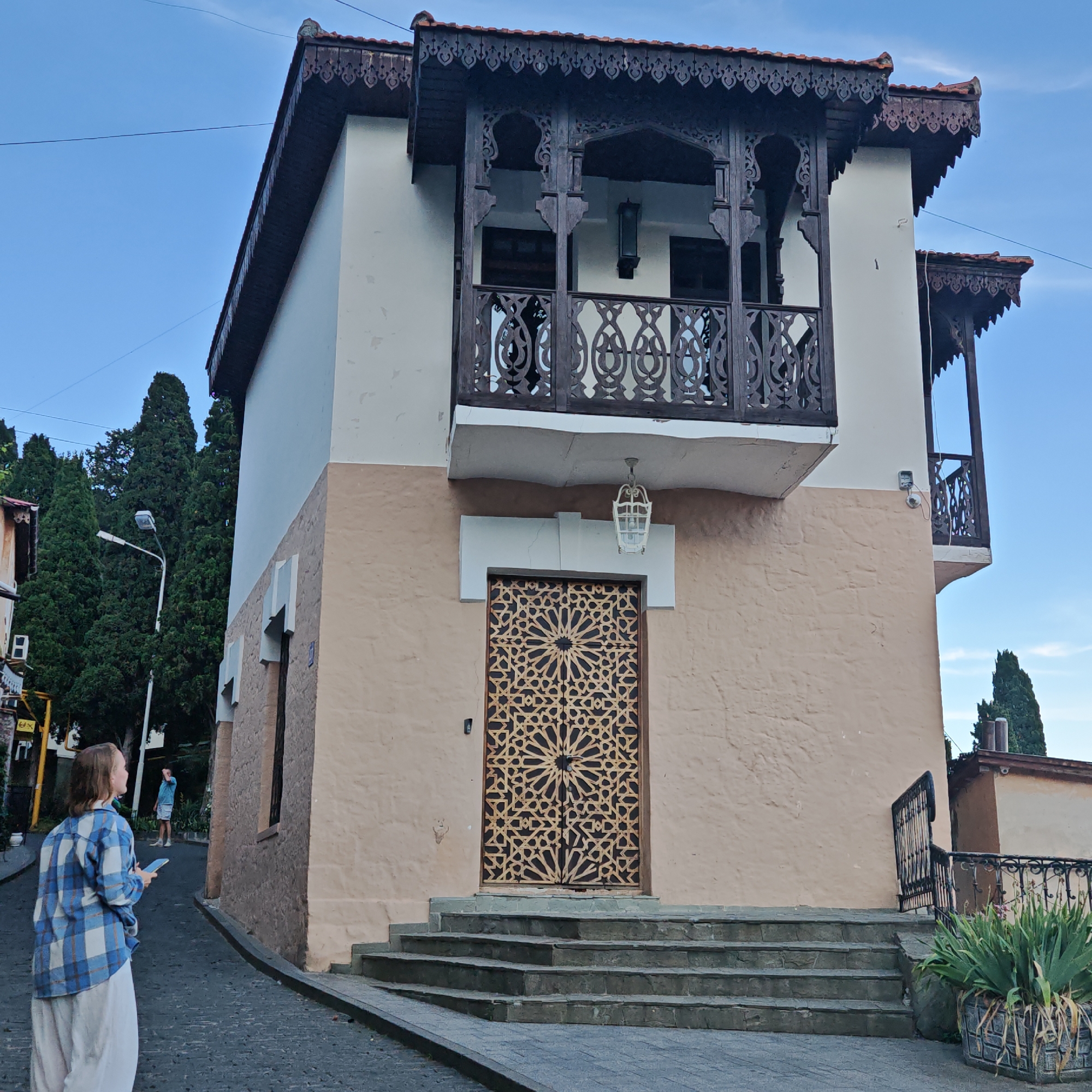 A young woman, wearing a blue and white plaid shirt and a long, light-colored skirt, stands on a cobblestone street, looking towards a building. She holds a phone in her right hand. Behind her, a man in a blue t-shirt and shorts stands further down the street. The building in the foreground is a two-story structure with a light beige lower half and a cream-colored upper half. It features ornate wooden balconies with intricate latticework and decorative eaves. A striking metal door with a geometric pattern is set into the lower level, accessed by a set of stone steps. A white lantern hangs above the door. The street is paved with dark cobblestones and slopes upwards. Tall, dark green cypress trees frame the building on the left. The sky is a clear, light blue, suggesting it is daytime, likely late afternoon or early evening based on the lighting. The overall scene appears to be in a historic, possibly Mediterranean or Eastern European setting, given the architectural style and the presence of cypress trees.