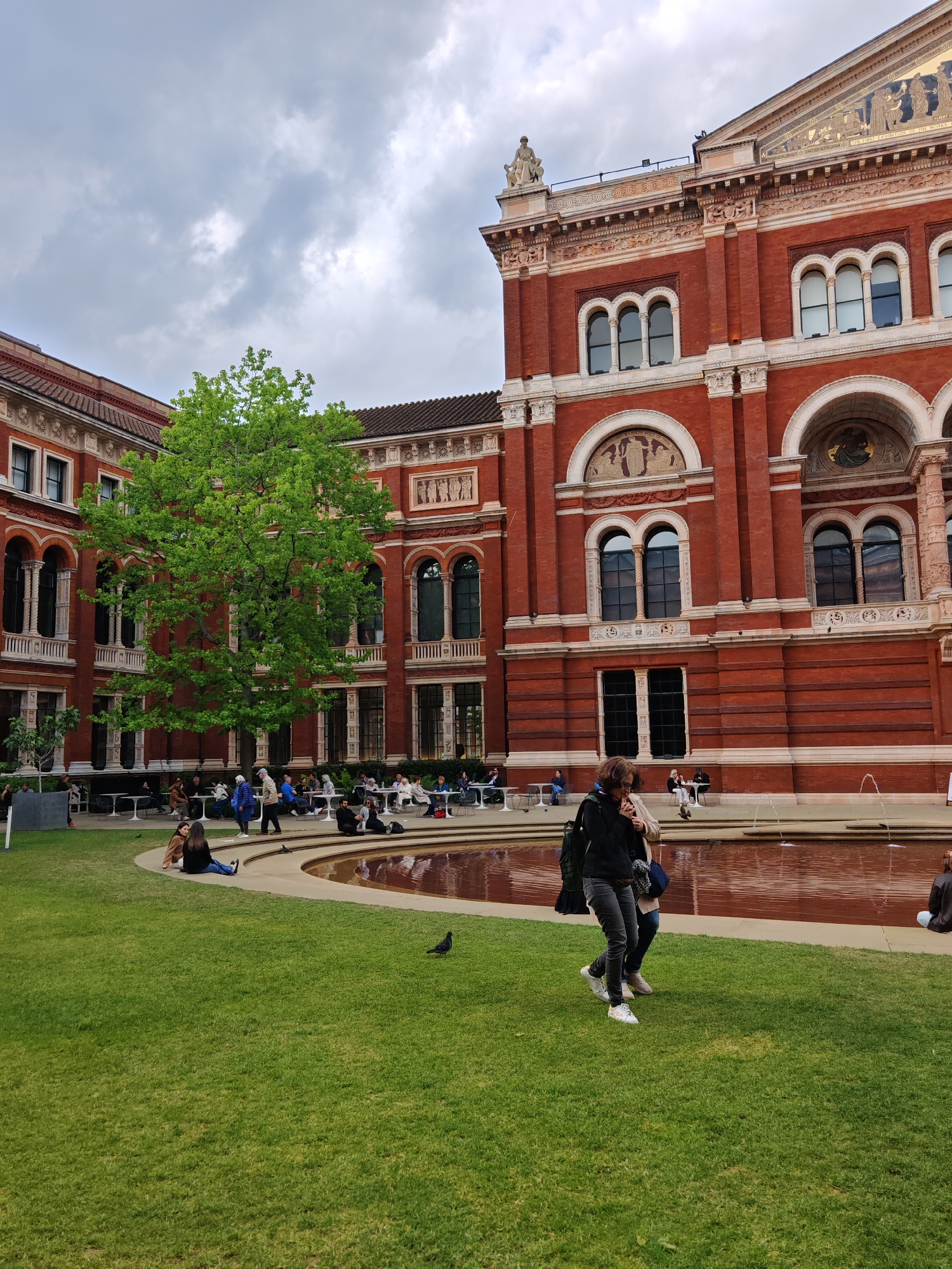 The image depicts a courtyard at the Victoria and Albert Museum in London, United Kingdom. The courtyard features a circular pond with water features. The architecture surrounding the courtyard is characterized by red brick buildings with ornate facades and arched windows. People are seen seated at tables, on the grass, and walking near the pond. A pigeon stands on the lawn in the foreground. The sky is overcast.