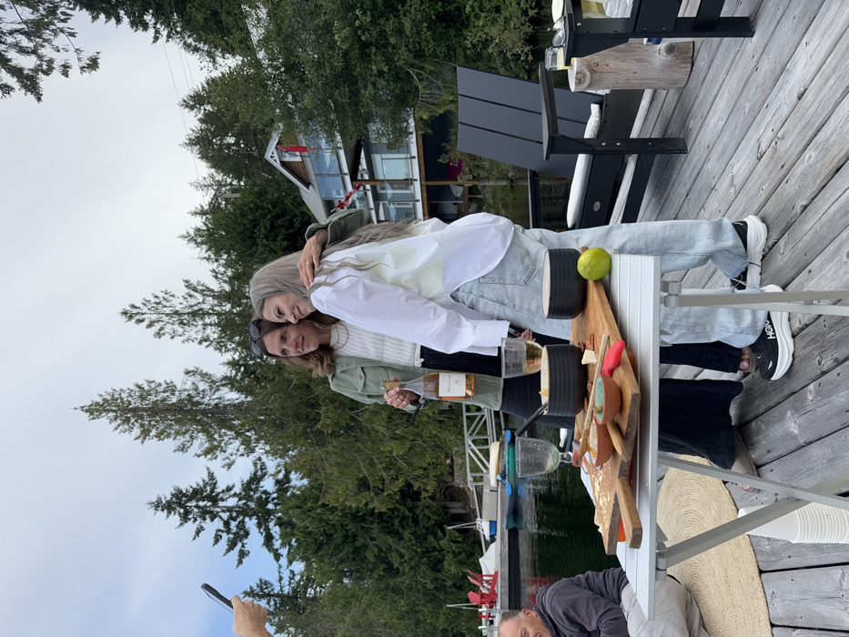 The image depicts an outdoor social gathering on a wooden dock at Area B (Shawnigan Lake), Canada, under an overcast sky. Two adult females are positioned centrally, one with brown hair wearing a green jacket over a white sweater and black pants, holding a bottle of rosé wine. The other, with long grey hair, wears a white collared shirt over light blue jeans and black and white Hoka shoes, with her left arm around the first woman. Both are smiling and looking towards the left. A partially visible hand holding a black smartphone is visible in the upper left corner, aimed towards the two women.
In the foreground, a silver collapsible table holds a light brown wooden serving board. The board features a variety of small bowls containing food items, a whole lime, and a red-handled knife. Two clear wine glasses are also present on the table, one containing a light-colored liquid. To the left of the table, a partially visible adult male with short dark hair is seated. A circular, light-brown woven mat is visible beneath the table, and a stack of white disposable cups rests on the dock surface.
In the background, calm, dark lake water extends behind the dock. Several small boats, including kayaks and paddleboards, are moored near the dock. Tall evergreen trees line the distant shore. A multi-level house with grey siding, brown accents, glass railings, and a prominent red Canadian flag hanging from a balcony is visible amongst the trees on the opposite shore or a nearby property. Two black Adirondack chairs and a wooden stump table are visible to the right of the central figures on the dock.