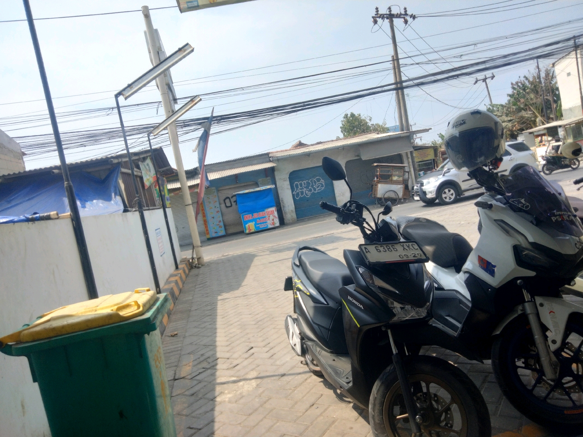 The image depicts a street scene in Pisangan Jaya, Indonesia. In the foreground, two scooters are parked on a brick-paved sidewalk. The scooter on the left is black with yellow accents and a license plate reading "A 6386 XKC 09-29". A helmet is placed on its handlebars. The scooter on the right is white and appears to be larger. Behind the scooters, a green trash bin with a yellow lid is visible.
The background features a row of buildings with corrugated metal roofs and storefronts. One storefront has a blue shutter with graffiti. A white car is parked in front of one of the buildings, and another scooter is in motion on the street. Power lines crisscross the sky, and a utility pole with multiple electrical lines stands on the right side of the image. The overall environment suggests a bustling urban or semi-urban area.