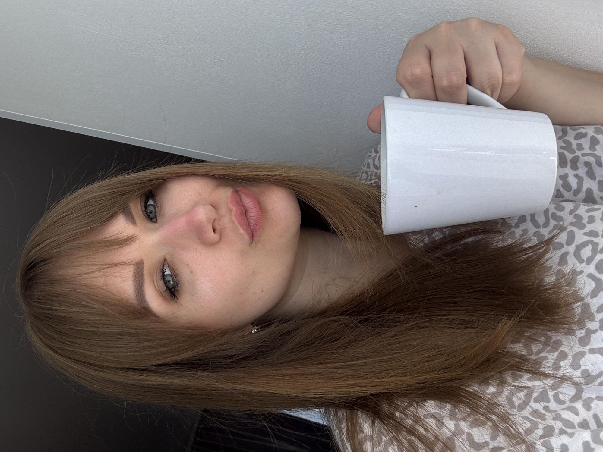 A woman with light brown hair and bangs is holding a white mug. She has green eyes and is wearing a patterned shirt. The background is a white wall with a dark area below. The image does not show any location that would indicate it was taken in the city of Yevpatoriya, Ukraine.
