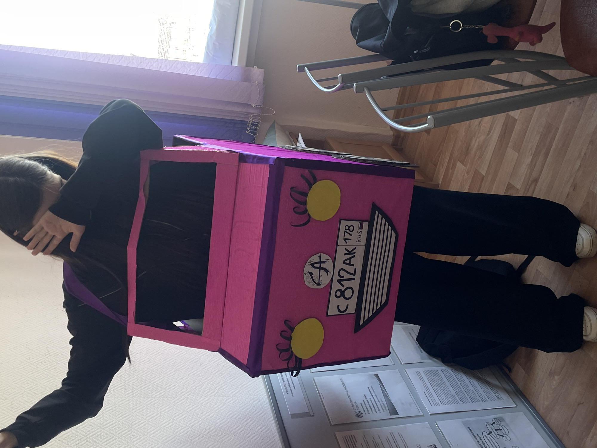 A person wearing a pink and purple cardboard bus costume is standing in what appears to be an indoor space, possibly a classroom or office. The costume has a Russian license plate on the back, indicating the city of Saint Petersburg, Russia (region code 178). The room has tiled flooring and is lit by natural light coming through a window. In the background, there's a metal-framed chair with a black bag on it. To the lower part of the picture there are papers hanging on the wall.