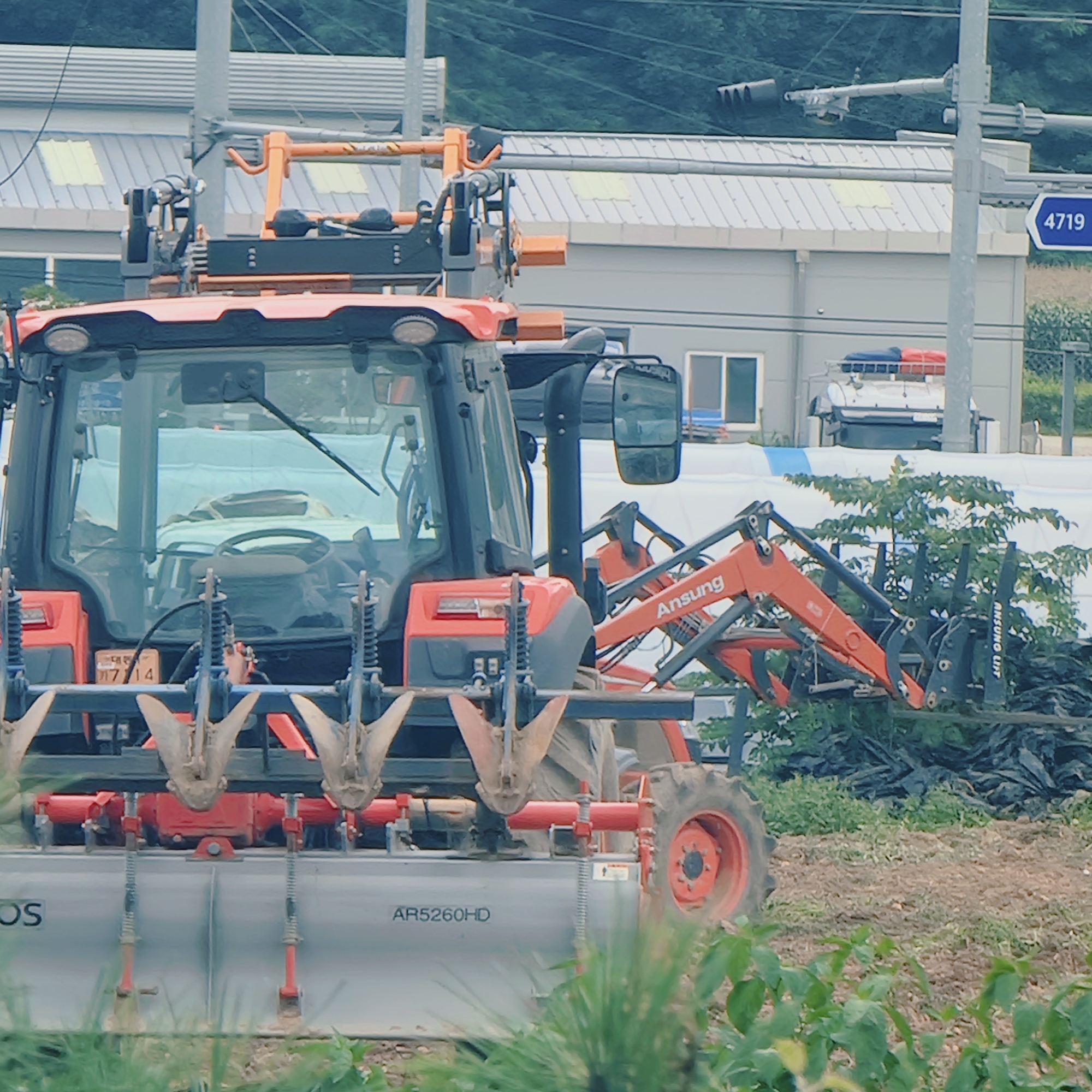 In Dunnae-myeon, South Korea, a bright orange tractor equipped with a plow attachment is positioned in a field. The tractor has a large, tinted windshield and a black cab. Attached to the front of the tractor is a row of metal plowshares. A hydraulic arm, also orange, with "Ansung" and "ANSUIG LIFT" visible on it, extends from the right side of the tractor, gripping a pile of black plastic sheeting. In the background, a light-colored building with a ribbed roof and a blue sign indicating "4719" is visible, along with a traffic light. The environment appears to be rural, with green foliage and a dirt field. There are no people clearly visible in the image.