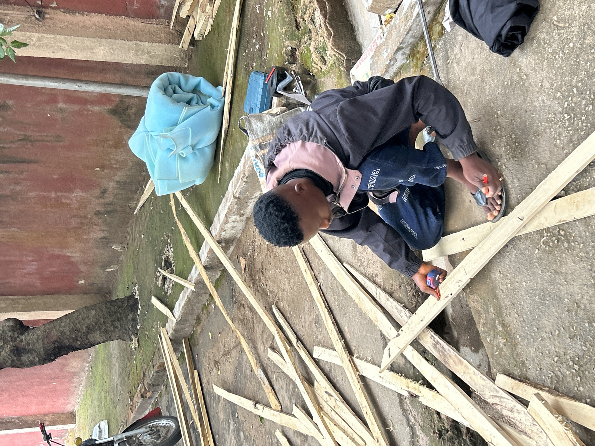 A person is kneeling on a concrete surface surrounded by numerous pieces of lumber, potentially in Jos, Nigeria. The individual is wearing a dark jacket, dark pants with a white logo, and sandals, and appears to be measuring or marking a piece of wood with a red object, possibly a pencil or marker. In the background, there are other items including a light blue cloth, a toolbox, and possibly a bag. A tree trunk is visible to the left, and a bicycle is partially visible at the bottom. The background also shows a red and brown wall and patches of green vegetation, suggesting an outdoor or semi-outdoor setting. The concrete surface is textured and shows signs of weathering.