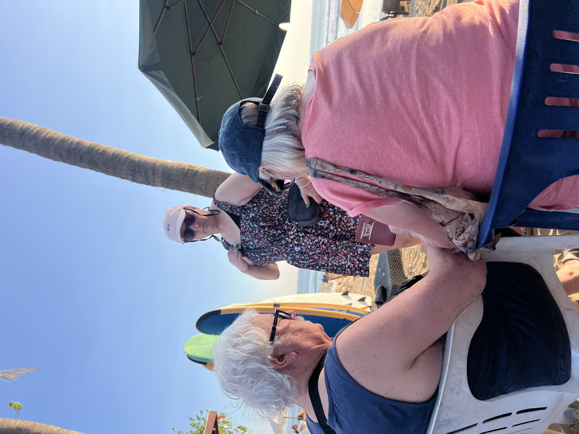 Three individuals are gathered on a sandy beach in Kuta, Indonesia, near a palm tree. The person in the foreground, on the left, is facing away from the camera and wearing a blue tank top and glasses. To their right, a person wearing a pink t-shirt and a blue baseball cap is also facing away from the camera. In the background, a third person in a floral dress and a white baseball cap stands with their back to the camera, facing the other two. Surfboards are visible behind the person on the left, and a green umbrella shades a seating area. The ocean and sky are in the background.