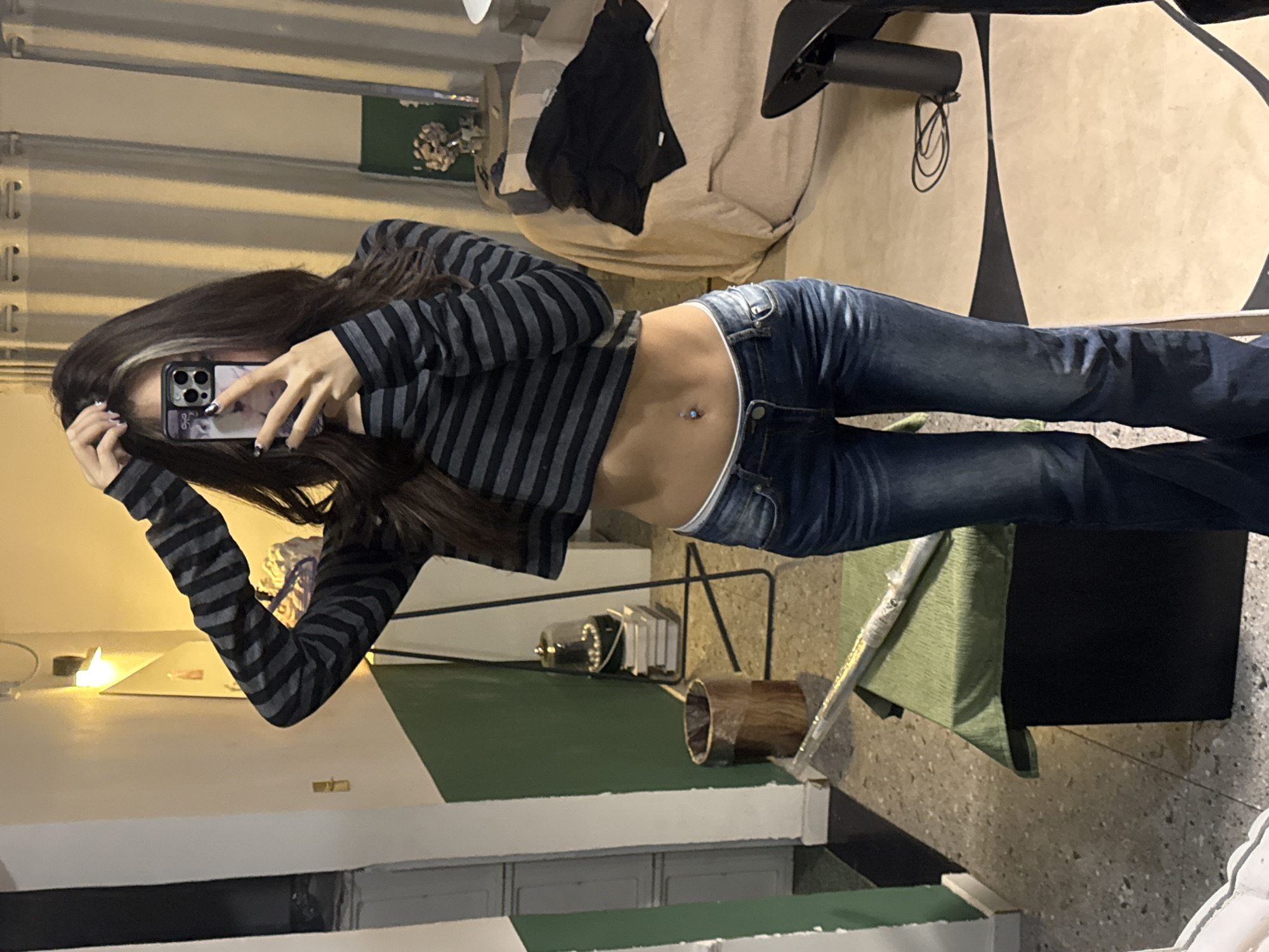An individual is depicted taking a mirror selfie indoors in Liangjiang, China. The subject, a person with long, dark brown hair featuring a lighter streak on the left side, wears a dark grey and black horizontally striped long-sleeve crop top, revealing a navel piercing with a light blue gem, and dark blue low-rise denim jeans exposing a white waistband. The subject holds a smartphone with a patterned case, obscuring their face, with both hands, one arm raised above the head. Fingernails are painted with a black and white design.
The background shows a room with a white wall, a dark green painted lower section, and a speckled light grey floor. A wall-mounted light fixture casts a warm glow on the upper left wall. Visible objects include a wooden cylindrical trash can, a stack of books topped with a glass cloche containing white spherical items, and a black metal stand. A dark rectangular object covered with a light green fabric has a silver-wrapped item resting diagonally on it in the lower left foreground. On the right side, light-colored curtains are visible, along with a beige upholstered chair or sofa draped with a black jacket. A small table or shelf with a floral arrangement is positioned in the upper right. The floor on the right features a light-colored rug with a dark, wavy pattern, alongside a black cylindrical object and a coiled black cable.