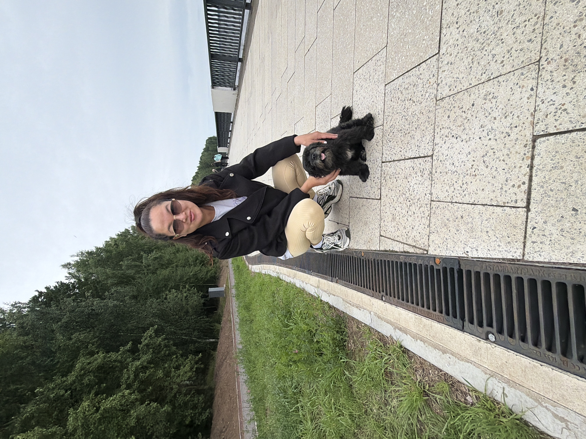 A young woman with long dark hair and sunglasses is kneeling on a paved walkway, petting a small black dog. She is wearing a black jacket over a white t-shirt and beige pants. The dog, with its tongue out, looks happy. The walkway is made of light-colored stone tiles with dark speckles. To the left of the walkway, there is a grassy area and a line of trees. Beyond the trees, the sky is overcast. To the right of the walkway, there is a metal grate, possibly a drain cover, running along the edge. In the background, there is a railing and a glimpse of more trees and a cloudy sky. The image appears to have been taken during the daytime. Some text is visible on the metal grate, but it is largely illegible. The overall scene suggests a casual outing with a pet in a park-like or public outdoor space.