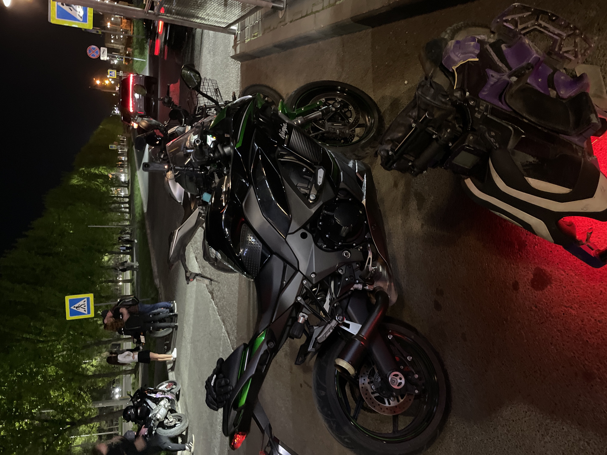 The image shows a street scene at night in Kazan, Russia. Several motorcycles are parked along the sidewalk. One black and green Kawasaki motorcycle dominates the foreground, next to a damaged purple and black motorcycle. Further down the sidewalk, people stand next to parked motorcycles. In the background, streetlights and traffic create a blurred backdrop. Road signs are visible above the parked cars. The pavement is partially illuminated by a red glow from a motorcycle.