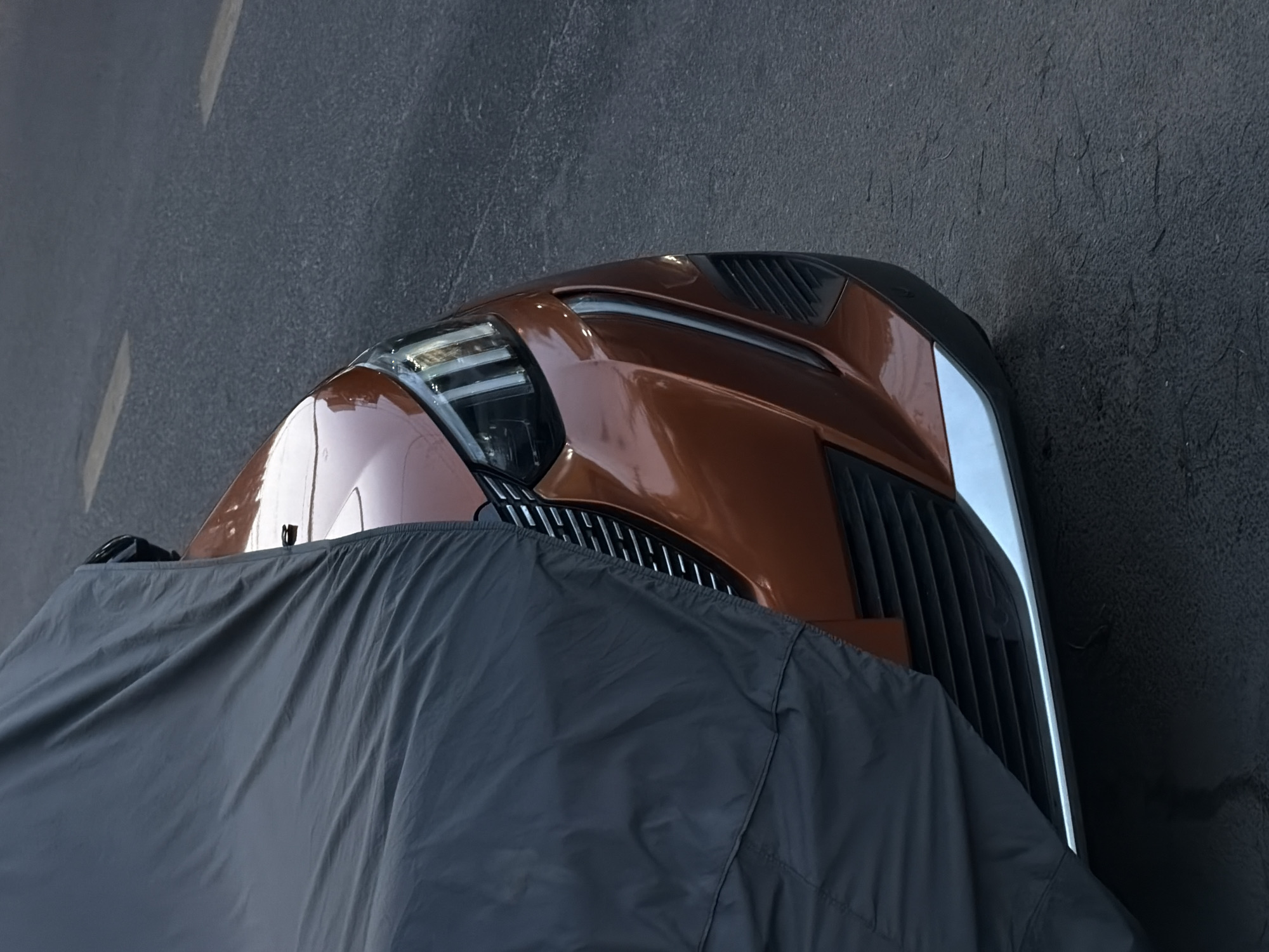 The image shows the front of a car partially covered by a gray protective cover. The car is a metallic bronze color and features a distinct headlight design with a white LED strip, a black grill, and vents on the hood. The car appears to be parked on a dark gray asphalt surface, with a blurred white stripe visible on the left side of the image, suggesting it is a street or parking area. The photo was taken in Đà Nẵng, Vietnam.