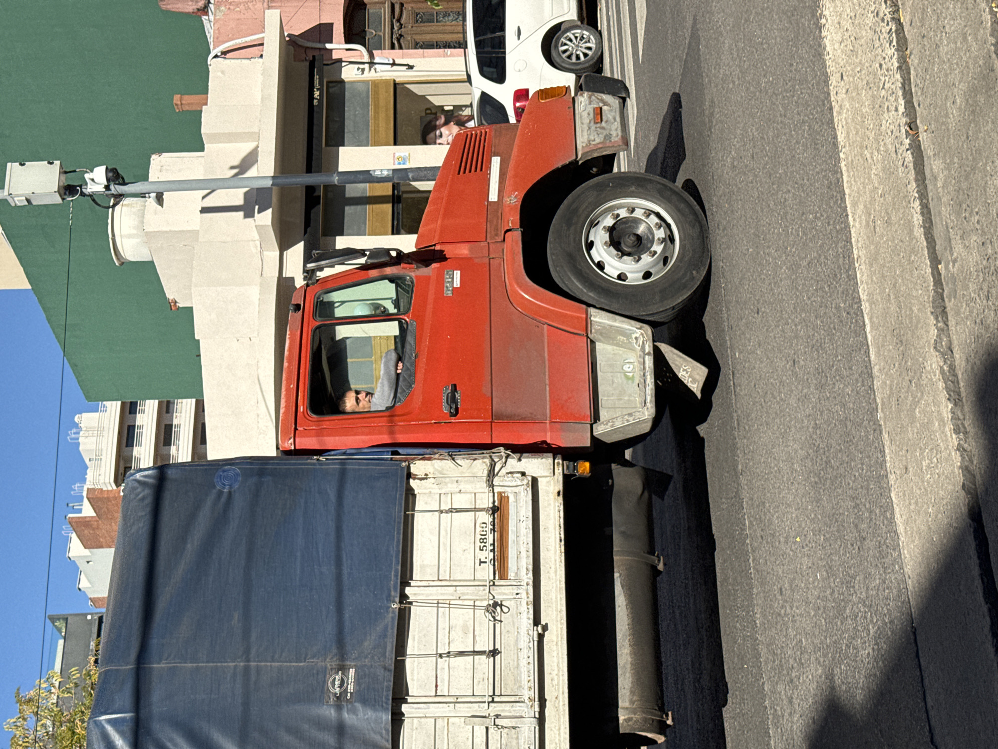 A red truck with a blue tarp-covered cargo bed is parked on the street in Buenos Aires, Argentina. The truck is oriented with the cab facing towards the upper left corner. The driver is visible in the cab. Buildings line the street. A white car is parked behind the truck, close to a building with architectural details and a security camera mounted on a pole. The street surface is paved with concrete, showing signs of wear.