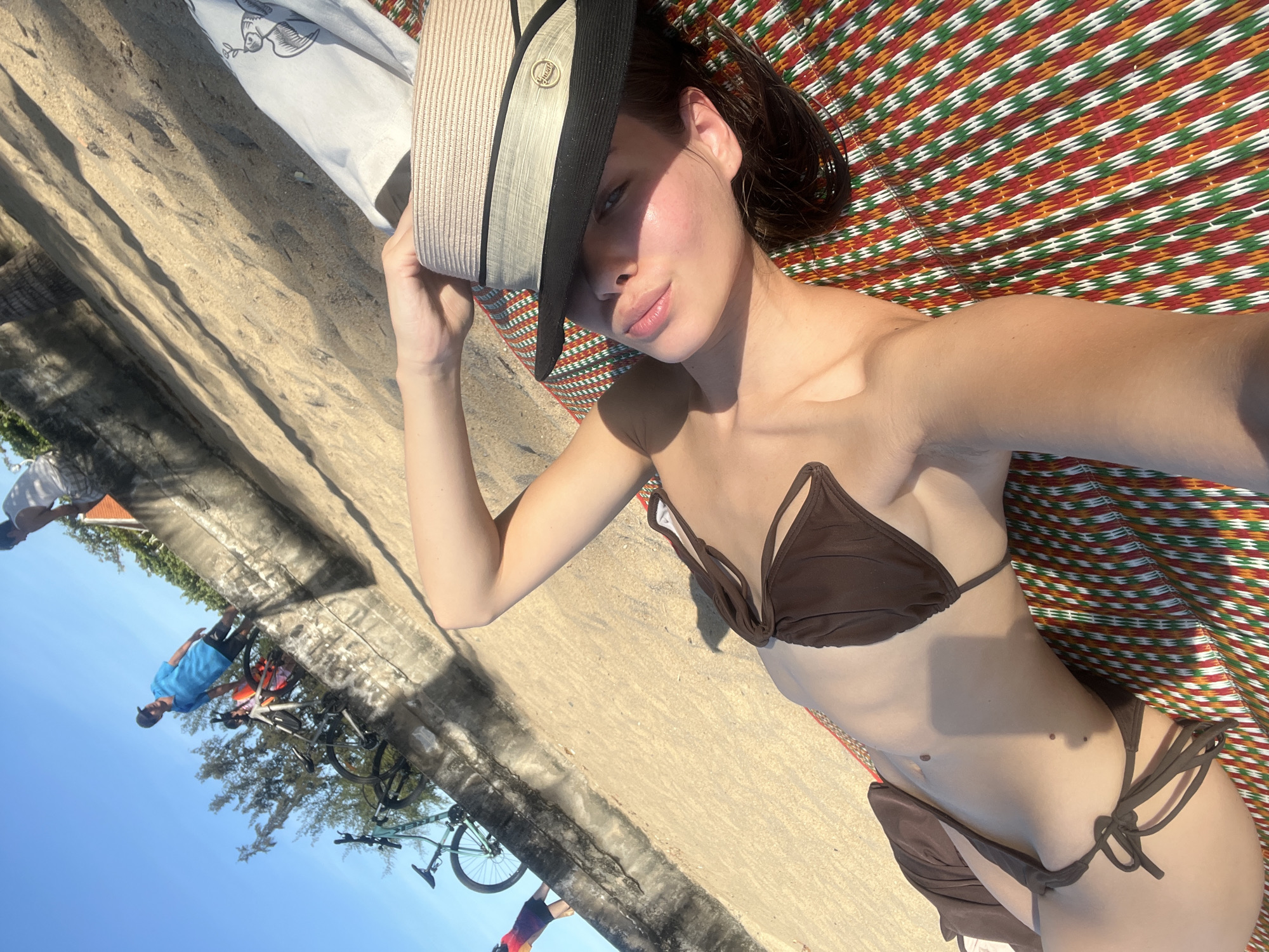 A woman wearing a brown bikini lies on a mat with a colorful geometric pattern, partially shaded by a straw hat with a dark brim that she holds. She is positioned diagonally in the frame, with her head near the top and her legs toward the bottom. The background transitions from a concrete area near the woman to an outdoor scene with people, bicycles, trees, and the sky. The photograph was taken in Nha Trang ward in the Khanh Hoa Province of Vietnam, as is evident from the outdoor scenery.