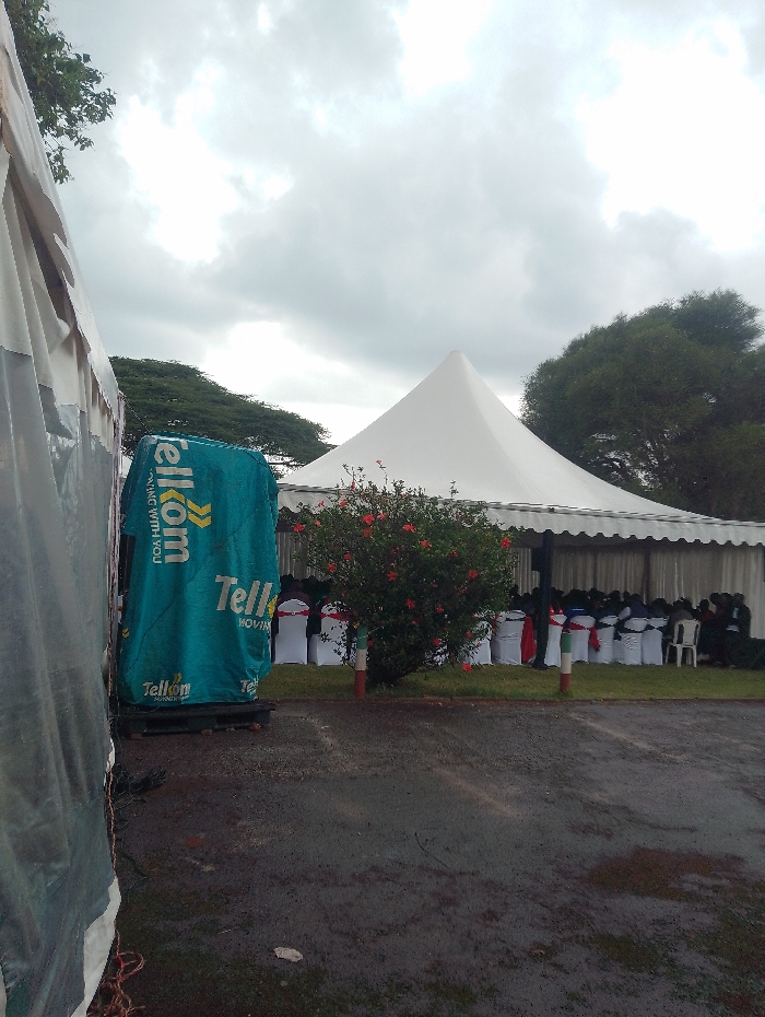 This image depicts an outdoor event venue in Gitaru, Kenya, set up under an overcast sky. The ground, a dark asphalt, appears wet, suggesting recent rainfall.
The scene is dominated by two white event tents. In the mid-ground, a large, rectangular tent features scalloped edges and provides shelter for numerous white chairs arranged in rows. Many of these chairs are decorated with red and blue sashes, hinting at a formal gathering or celebration. Behind this main structure, a smaller, conical white tent stands.
To the left, a tall, green object covered in a branded banner prominently displays the "Telkom" logo and the text "MOVING WITH YOU," indicating corporate involvement, likely a sponsorship or a company event. A vibrant bush with bright red flowers is situated in front of the main tent, flanked by two short, striped poles (red, white, and green).
In the background, lush green trees contribute to the outdoor setting. The visible seating arrangement suggests an organized gathering or ceremony, possibly a community or corporate event given the Telkom branding. The overall atmosphere is one of anticipation for or during an event, under grey, cloudy conditions.