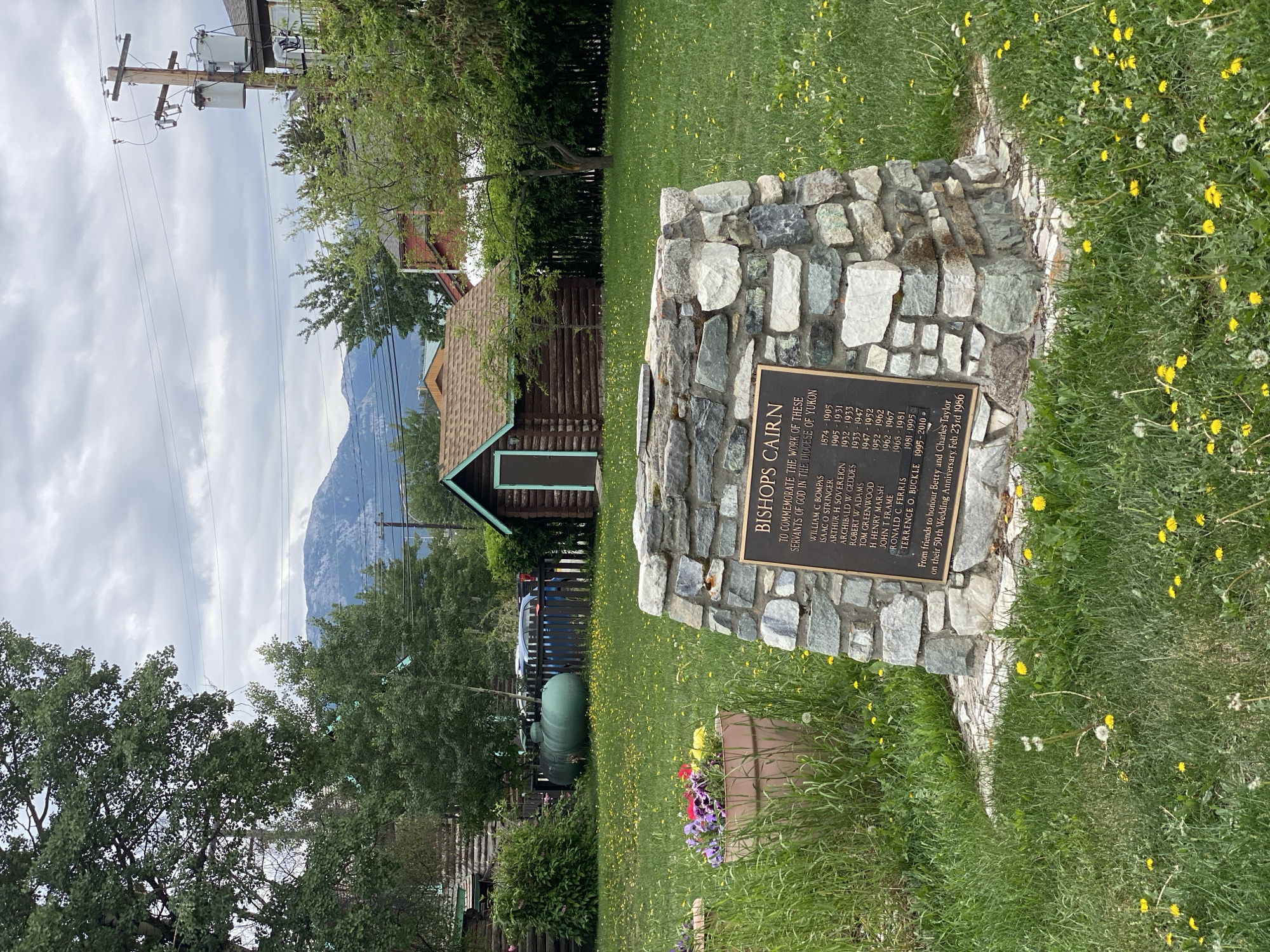 The image depicts an outdoor scene in Whitehorse, Canada, focused on a stone monument in a grassy area dotted with dandelions. The monument, constructed from various stones, features a bronze plaque inscribed with text. The plaque's title reads "BISHOPS CAIRN" and lists several names with corresponding years, seemingly indicating a historical or commemorative purpose. Below this list, it states, "From friends to honour Betty and Charles Taylor on their 50th Wedding Anniversary, Feb 23rd 1956."
In the background, a rustic wooden shed with a brown roof is visible, set against a backdrop of lush green trees and a chain-link fence. Beyond the fence, two large green tanks are situated, and a blue vehicle is parked behind them. A distant mountain range, partially obscured by clouds, forms the horizon. The sky is overcast, contributing to the soft, diffused lighting of the scene. The overall impression is one of a peaceful, possibly historic, location within the city, marked by the monument and its surrounding natural elements.
