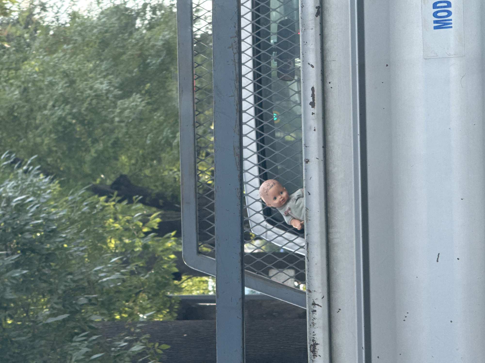 The image shows a doll looking out from behind a metal grate on the side of a truck. The truck is silver with a vertical seam along the side. A sign reading "MOD" is partially visible at the top. The background features green trees and a glimpse of a vehicle through the grate. The doll is light-skinned with dark eyes and a grey shirt. The location is Puebla, Mexico.