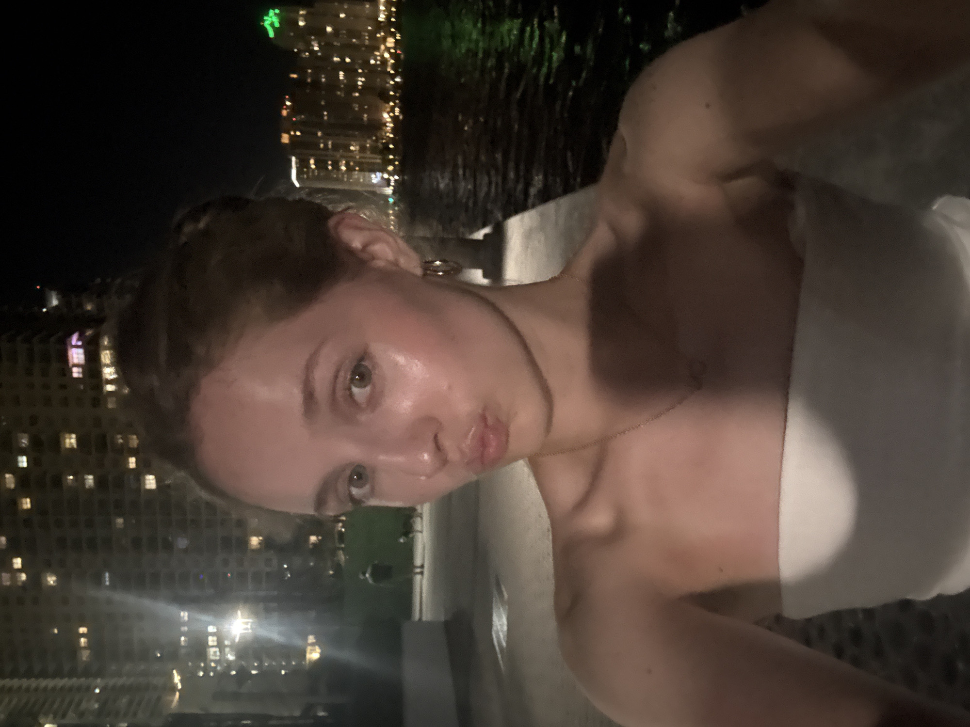 A young Caucasian woman with blonde hair pulled up into a bun is taking a selfie at night. She is wearing a white strapless top and a delicate necklace. She is outdoors, with a body of water to her right and a city skyline with illuminated buildings in the background. The woman is in the foreground, and her face is turned towards the camera with her lips pursed. In the background, a man and a dog are visible on a paved pathway. The environment appears to be a waterfront promenade in Miami, USA, likely near the Biscayne Bay area, given the cityscape and body of water. The lighting is dim, with the city lights and some streetlights providing illumination.