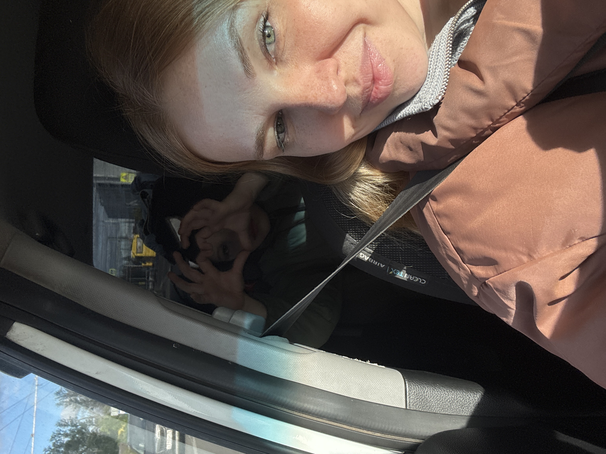 The image depicts a selfie taken inside a car. A woman with fair skin, light hair, and green eyes is visible in the upper portion of the frame. She is wearing a jacket with a gray collar. A seatbelt crosses her body.
In the background, a reflection in the car window shows another person. The reflection is obscured. Outside the car, a yellow construction vehicle is visible. The surroundings appear to be an urban area with trees visible. Rusanovo, Russia cannot be determined.