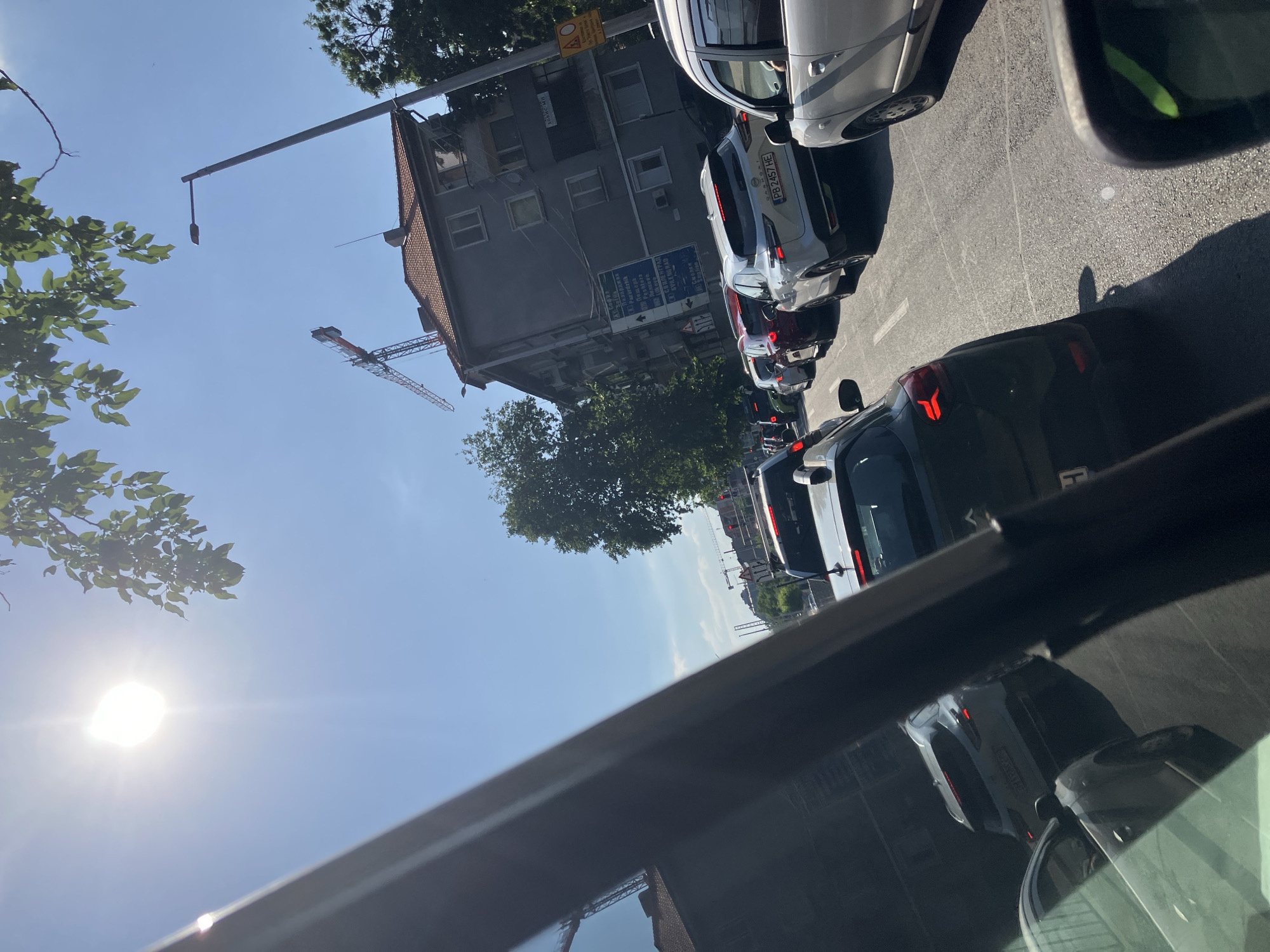 The image, taken from inside a car, shows a traffic jam on a city street in Plovdiv, Bulgaria, based on the Cyrillic signage on a building in the background. The camera is pointed upwards, capturing a view of the cars ahead, a multi-story building, a construction crane, and the sky. Bright sunlight is visible. The sign lists Pazardzhik, Peshtera and other destinations.