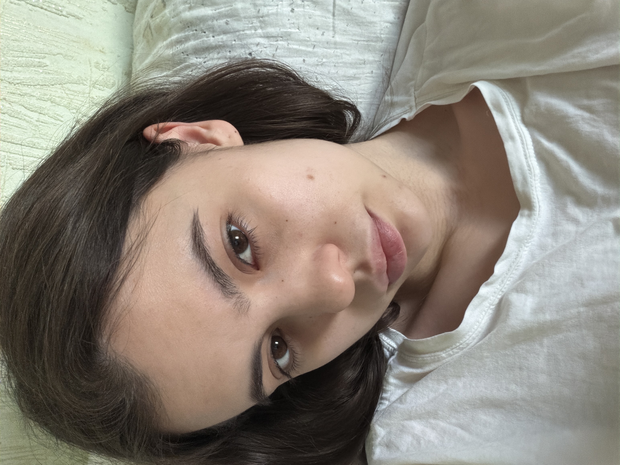 A young woman with dark brown hair lies on a light-colored pillow and white t-shirt. She is looking directly at the camera with her brown eyes. The location is not discernible from the image.
