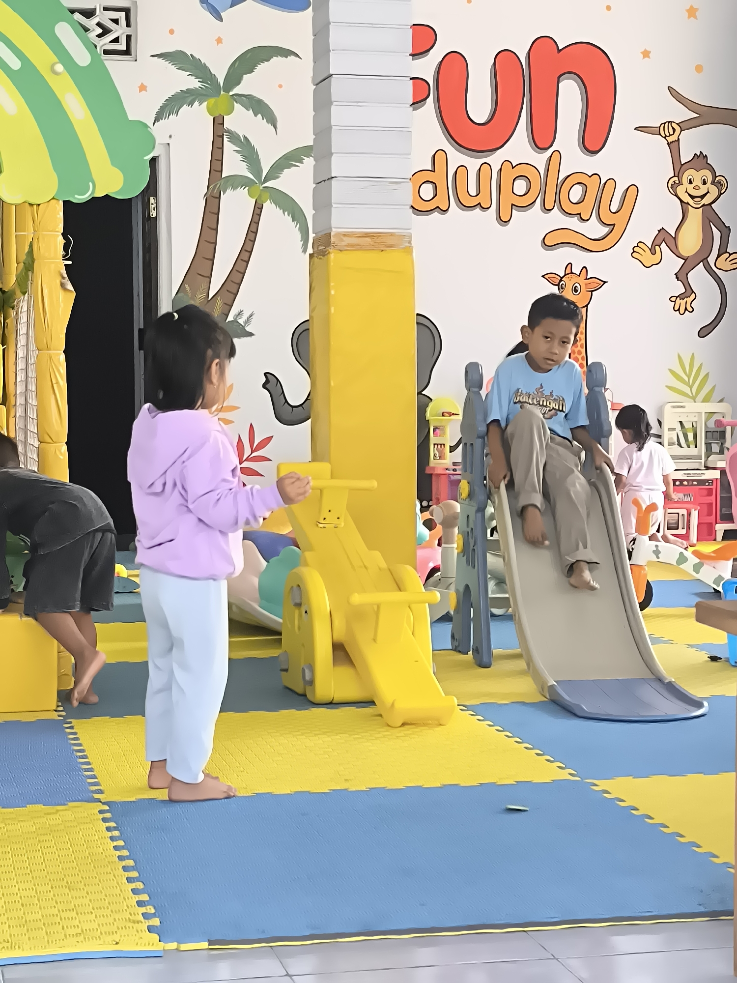This image depicts a playful indoor environment, possibly a children's play area, featuring vibrant decorations and play equipment. A mural on the wall displays tropical elements like palm trees, a monkey hanging from a branch, and the words "Fun duplay" in large, colorful lettering. In the foreground, a young boy is seated on a plastic slide, looking towards the camera with a neutral expression. He is wearing a light blue t-shirt and brown pants. Beside the slide, a young girl in a lavender hoodie and white pants stands near a yellow play structure, holding onto a handle. In the background, other children and various toy kitchens and play appliances are visible, suggesting a space designed for children's entertainment. The floor is covered with interlocking foam mats in yellow and blue. The overall impression is one of a bright and active recreational space. The text overlay "Dagangan, Indonesia" suggests the location of this scene.