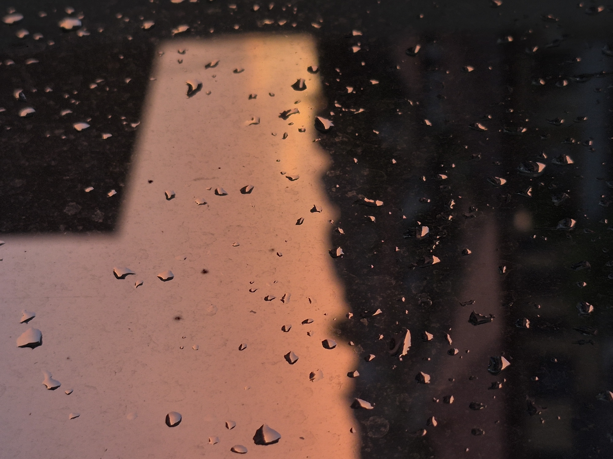 The image depicts a close-up view of a windowpane covered in raindrops, with a blurry sunset visible in the background. The sky transitions from a soft peach color at the horizon to a deeper, dusky orange higher up. The silhouette of a treeline or buildings is discernible against the colorful sky. The raindrops on the glass create distorted reflections and magnify the colors of the sunset, adding texture and depth to the scene. The overall impression is one of a quiet, contemplative moment, possibly at dusk. The location in Popovo, Bulgaria, is not visually apparent from the image itself.