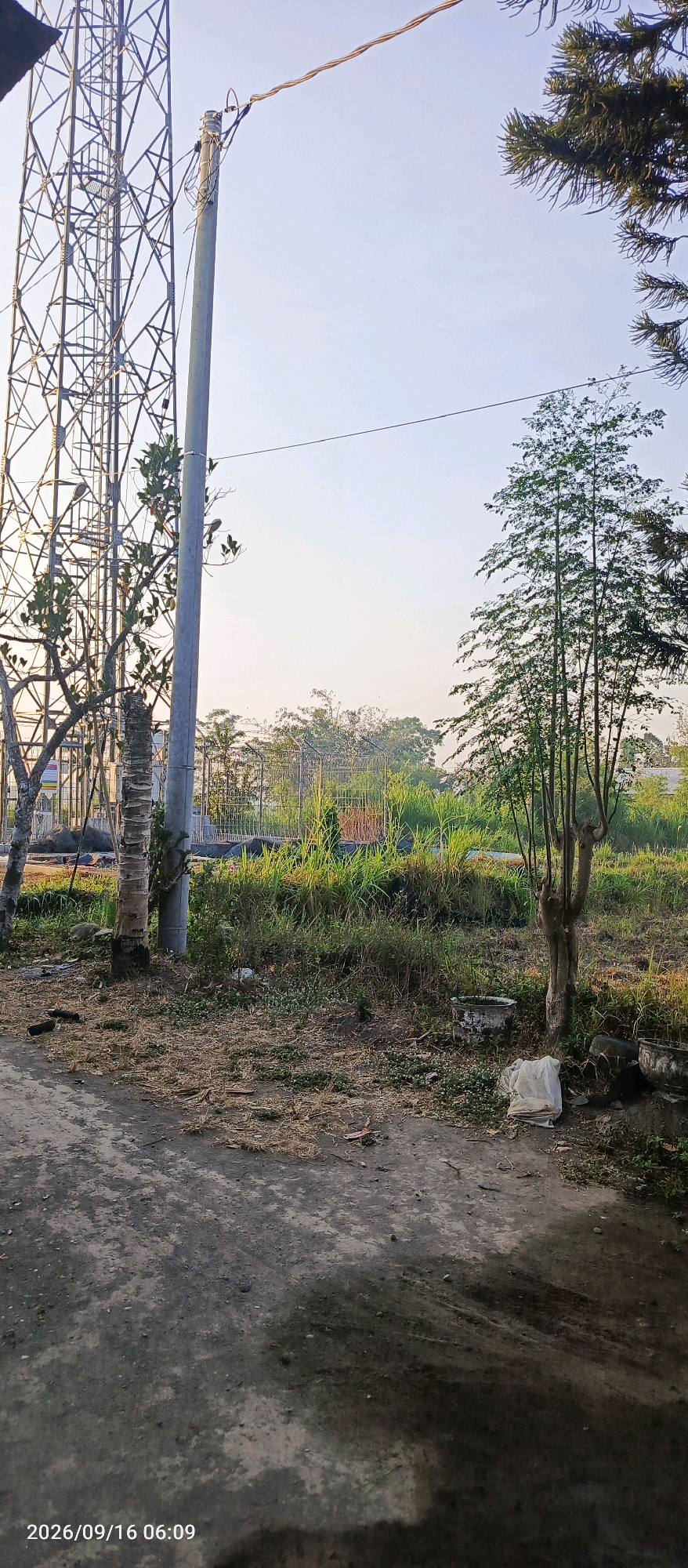 The image shows a telecommunications tower and a utility pole, with overhead wires connecting them. The tower, a lattice structure, is on the left side of the image, while the pole is in the center. The background is a landscape with trees, tall grass, and some fencing structures. There is also a dirt or gravel path in the foreground. The sky is clear and pale blue, suggesting it is daytime. The overall scene appears to be an outdoor area, possibly on the outskirts of a town or in a rural setting. A timestamp in the lower left corner indicates "2026/09/16 06:09". The location is specified as Karangsono, Indonesia. There are no people visible in the image.
