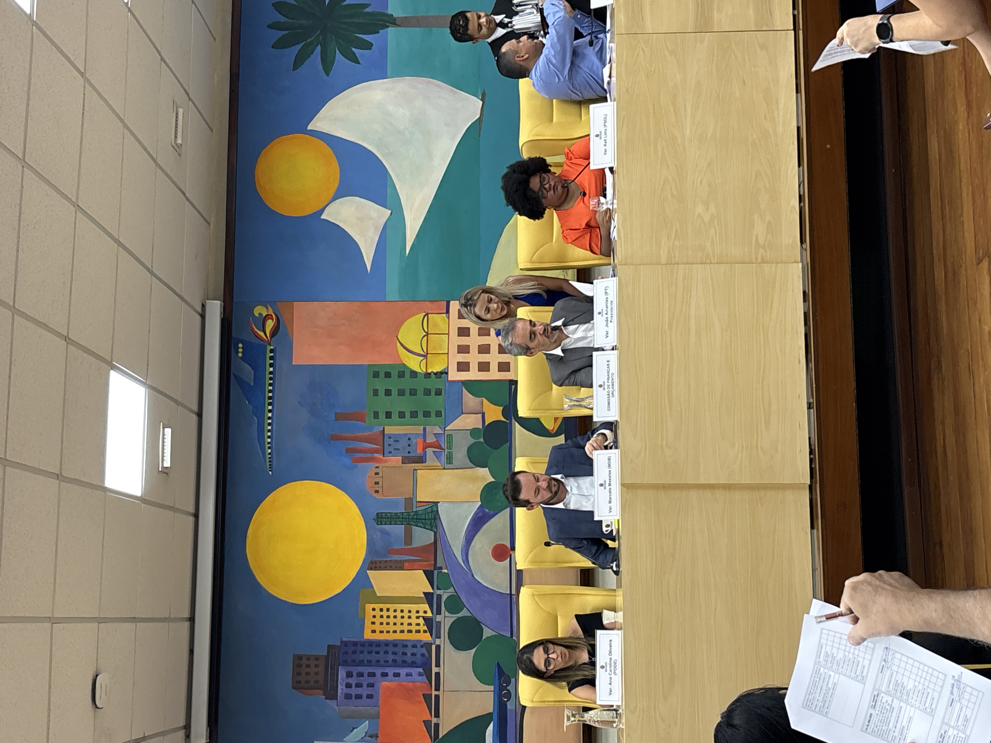 The image shows a meeting taking place in a room. There are several people seated at a long table. Nameplates are in front of each person, identifying some individuals as Ver. Ana Carolina Oliveira, Ver. Marcelo Messias, Ver. João Ananias, and Ver Kelt Lima. Some individuals are wearing business attire.
Behind the table is a large mural depicting a cityscape with a harbor and a sailboat under a yellow sun. The mural likely represents the city of São Paulo, Brazil, where the meeting is located.
The room itself appears to be a formal meeting space, with a panelled wall along the side. Some people are holding papers.