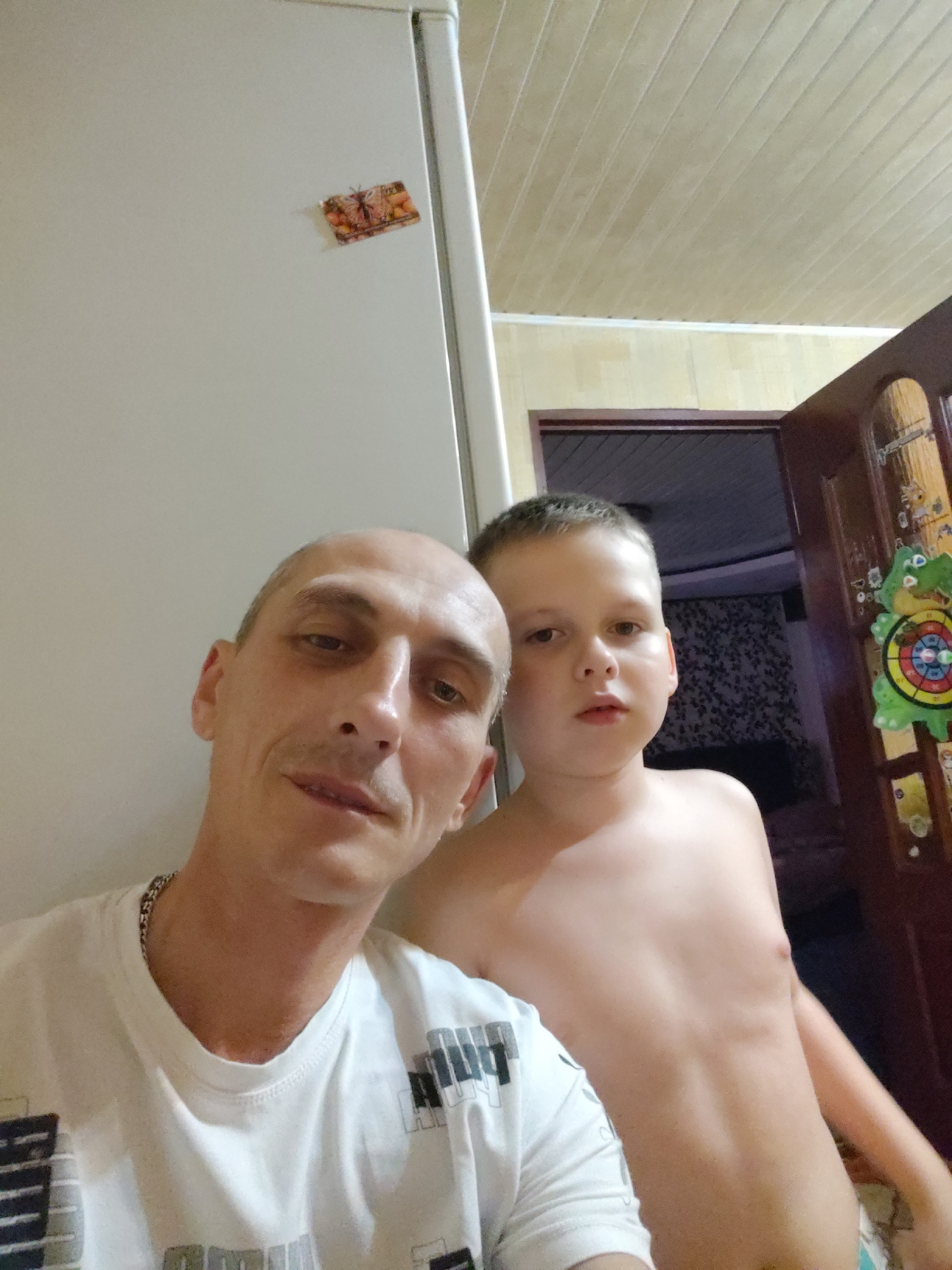 The image is a selfie taken indoors. Two subjects are visible: a man in the foreground and a young boy behind him. The man is bald and wearing a white t-shirt with a silver chain. He is looking at the camera with a neutral expression. The boy is shirtless and has short, light brown hair. He is also looking towards the camera with a slightly curious expression.
In the background, a refrigerator is partially visible on the left. A decorative magnet with an image of a butterfly is attached to the refrigerator door. To the right, a wooden door is visible, adorned with several colorful decorations, including a frog-shaped target. The room appears to be a home, possibly a kitchen or hallway, with a wood-paneled ceiling. The lighting suggests it is daytime. The overall mood is casual and familial. No visible text is discernible in the image beyond what might be on the t-shirt pattern.