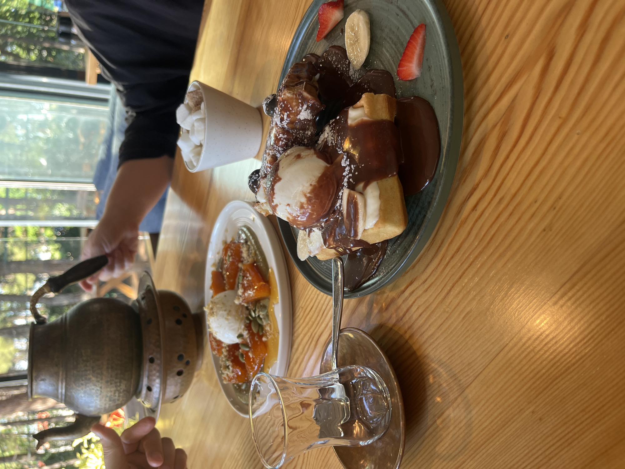 This daytime image captures an inviting dining scene at a cafe or restaurant in Kaspiysk, Russia. The wooden table, with its prominent diagonal grain, is laden with decadent desserts and traditional beverage items.
The central subject is a rich dessert served on a dark green plate. It features churros or similar fried dough pastries, heavily coated in glistening chocolate sauce, topped with a scoop of melting vanilla ice cream, and garnished with fresh slices of strawberry and banana. A light dusting of powdered sugar adds to its appeal. Beside this, a white ceramic cup overflows with small, white marshmallows, likely intended for a hot beverage.
To the left, a traditional, hammered copper or brass kettle/samovar with a dark handle sits on a stand, suggesting the serving of tea or coffee. Nearby, a clear, fluted glass cup, characteristic of Turkish tea glasses, rests on a saucer with a spoon. Another dish on an oval white plate holds what appears to be stewed apricots or peaches, crowned with a scoop of ice cream or clotted cream and sprinkled with pumpkin seeds, offering a fruity counterpoint to the chocolate-rich dessert.
Partially visible hands and arms of two individuals indicate people are actively engaged in the meal. The background reveals blurred greenery outside a large window, suggesting an outdoor view and natural lighting. The scene conveys a sense of indulgence and a leisurely dining experience. No text is visible in the image.