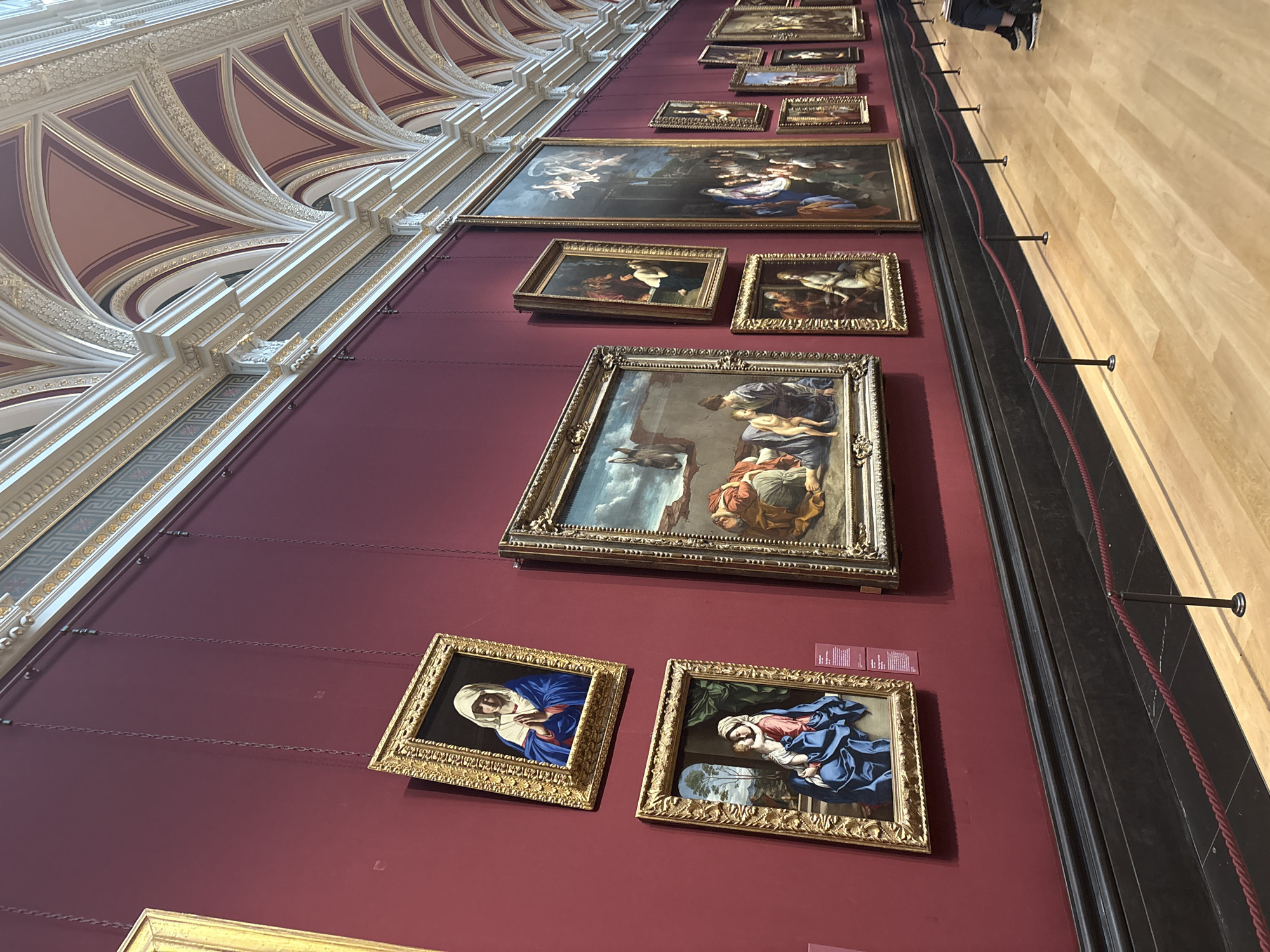 The image shows a wall in the National Gallery, London, United Kingdom. The wall is painted a deep red and is adorned with numerous framed paintings, primarily depicting religious and mythological scenes. The paintings vary in size and are hung in a somewhat haphazard arrangement. The architecture of the building is visible at the top left, showcasing an elaborate ceiling with ornate detailing and gold accents. A varnished wooden floor and crowd-control stanchions are visible to the right. A person's legs are seen in the background.