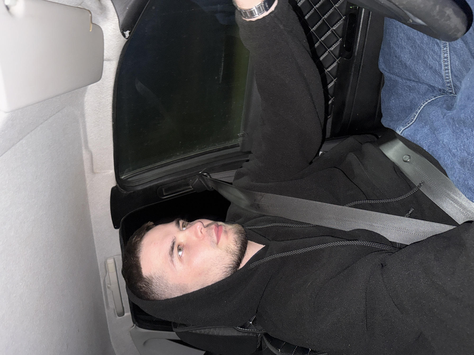 A man is sitting in the driver's seat of a car at night. He is wearing a black hooded sweatshirt and blue jeans. The man is looking to his right, and his left arm is extended, resting on the steering wheel. A seatbelt is fastened across his chest. The interior of the car is visible, including the dashboard and door panel. The windows are dark, and the outside environment is not clearly discernible, but appears to be a dark, potentially rural or suburban area. The image was taken in the city of Kondratovo, Russia.