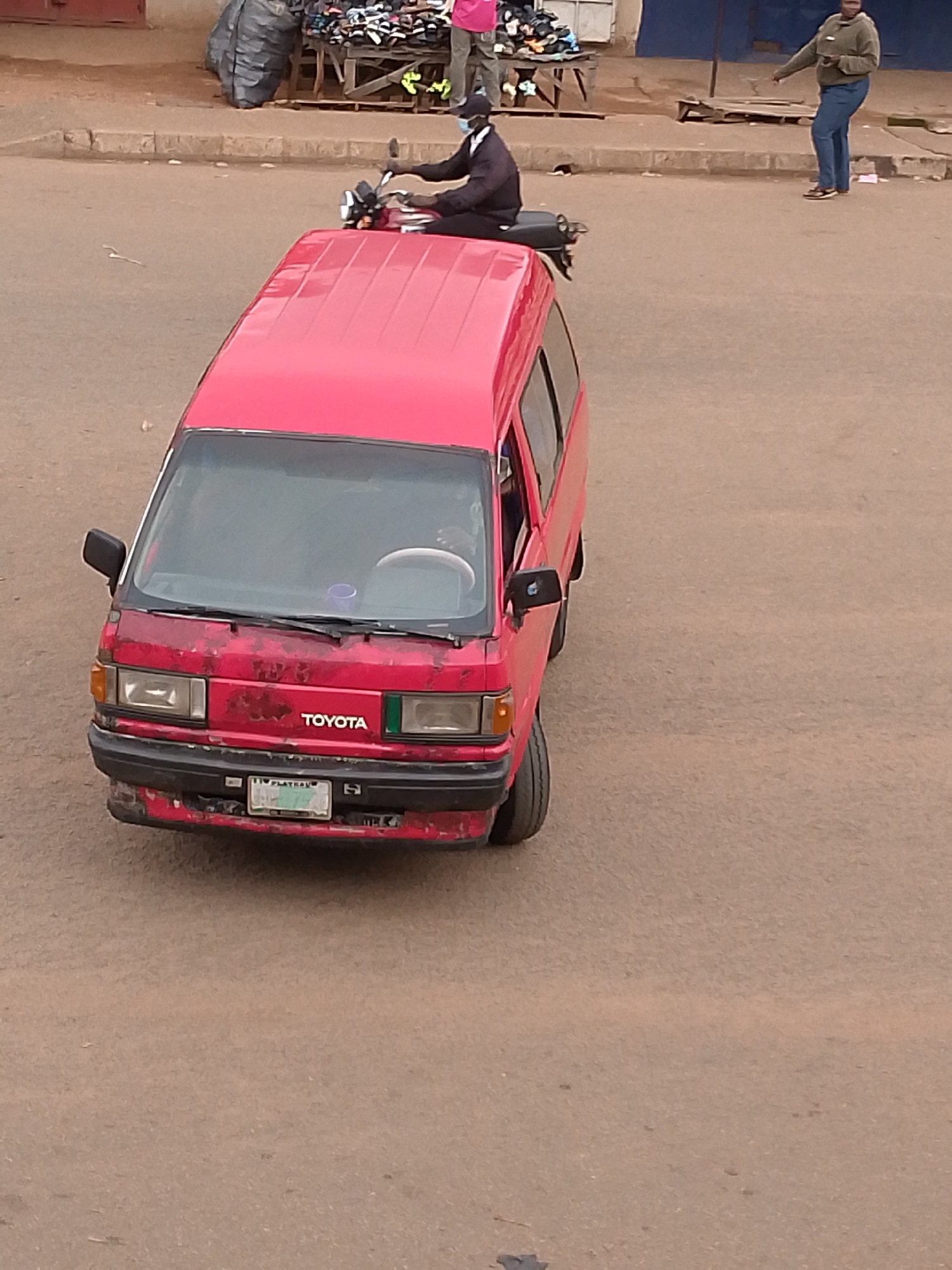 A red Toyota Hiace van, marked with "TOYOTA" on its front, is parked on a dirt road in Bukuru, Nigeria. The van shows signs of wear, with chipped paint on its front bumper and body. A license plate reading "VTS 4638" is visible on the front bumper. To the left of the van, a person wearing a black jacket and mask rides a red motorcycle. In the background, a stall displays various items, including shoes and bags, and a large black tarp is visible. Further in the background, two individuals are present; one wearing a green hoodie and blue jeans, and another partially visible person wearing a light-colored top and green pants. The ground is dusty, indicating a dry, unpaved area.