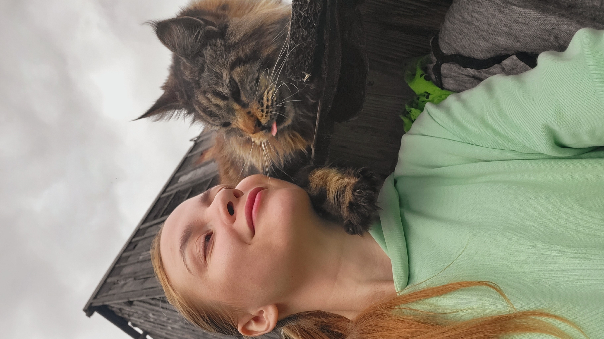 A young woman with light reddish-brown hair, wearing a mint green hooded sweatshirt, smiles with closed eyes as a large, long-haired tabby cat rests its head and one paw on her left shoulder. The cat, exhibiting prominent ear tufts, has its tongue slightly extended and appears relaxed. They are positioned outdoors in front of a weathered, dark wooden building, visible from a low angle against an overcast, light grey sky. The scene conveys an affectionate interaction between the subjects.