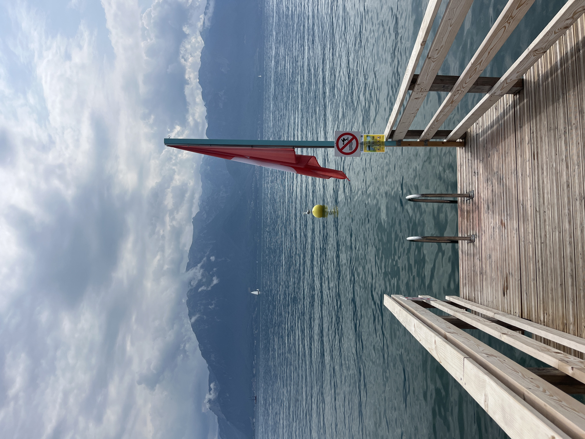 The image depicts a wooden dock extending into a tranquil body of water, likely Lake Geneva, with a backdrop of majestic mountains, characteristic of Vevey, Switzerland. A Swiss flag, predominantly red with a white cross, is unfurled on a pole to the right of the dock. Adjacent to the flag, a circular sign with a red border prohibits diving, indicated by a silhouette of a person diving into water with a red line through it, accompanied by French text. A yellow sign with additional text is mounted below it. A yellow buoy floats in the water near a lone seagull perched on it. In the distance, sailboats glide across the water, and a ship is visible further to the left. The sky is overcast with dramatic clouds, casting a subdued light on the scene. The dock itself is constructed of light-colored wooden planks and features metal handrails leading down to the water, suggesting a swimming or boating area. The water appears calm with gentle ripples, reflecting the cloudy sky and distant mountains.