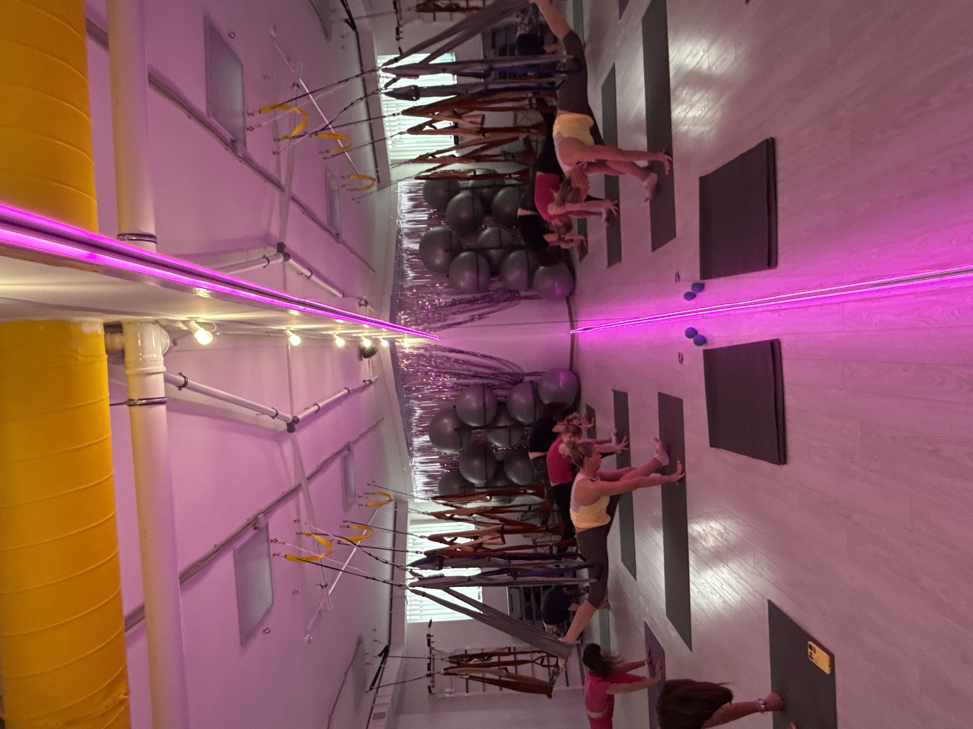 In an indoor fitness studio, a group of women participate in a stretching class. The room is illuminated with pink lighting and features exercise equipment such as suspension straps and exercise balls. The walls are painted in a light color, and yoga mats are placed on the floor. A bright yellow vertical pipe is visible on the left side of the image. The studio appears to be mirrored, with the scene reflected along a horizontal axis, though it is unknown if this is the intended location in Chaikovsky, Russia.