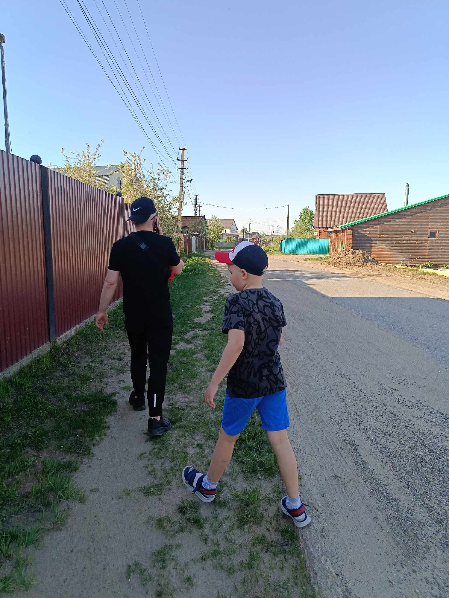 Two people, an adult and a child, are walking on a dirt road. The adult, wearing a black t-shirt, black pants, black shoes, and a black baseball cap with a white logo, appears to be talking on a cell phone. The child, wearing a patterned t-shirt, blue shorts, and sneakers, is walking slightly ahead. A brown corrugated metal fence borders the left side of the road, while houses and utility poles are visible on the right. The scene appears to be in a rural or semi-rural area with a clear sky. The location is Chaikovsky, Russia.