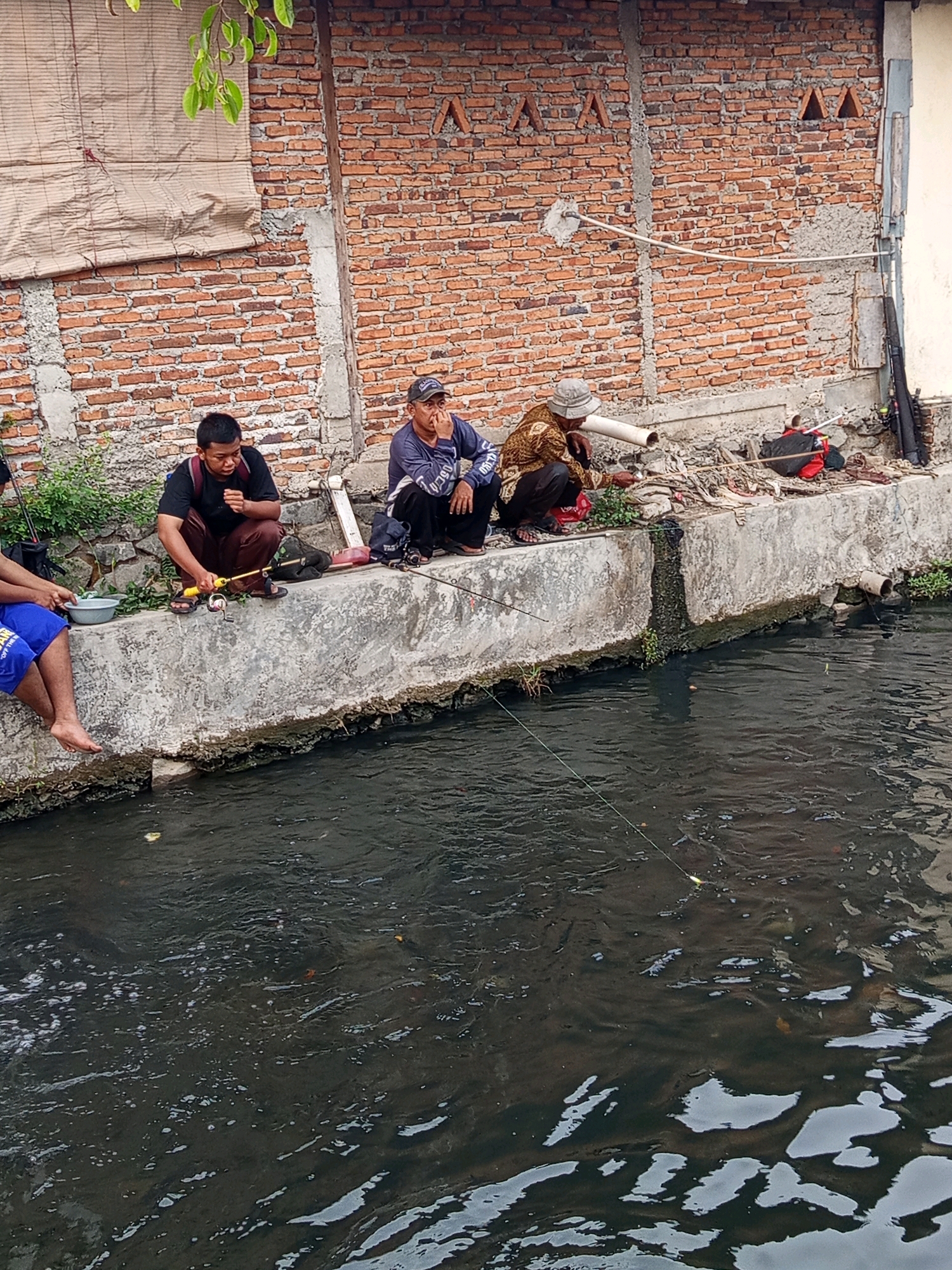 The image depicts a group of men fishing in a murky canal that runs alongside a brick wall in Karawang, Indonesia. Three men are prominently seated on the concrete edge of the canal. The man on the left wears a black t-shirt and brown trousers, holding a yellow fishing rod. The man in the middle is dressed in a blue long-sleeved shirt and dark trousers, wearing a black baseball cap and holding his hand to his chin. The man on the right is wearing a patterned long-sleeved shirt and dark trousers, with a light-colored hat, and is also fishing. Further to the left, another person is partially visible, wearing blue shorts. The environment appears to be urban, with the brick wall forming a backdrop, possibly part of a residential or industrial area. Debris and some greenery are scattered along the canal edge. The water is dark and appears polluted, with some ripples and reflections on its surface. Fishing lines extend into the water from the men's positions. A rifle-like object leans against the wall on the far right, near some fishing equipment.