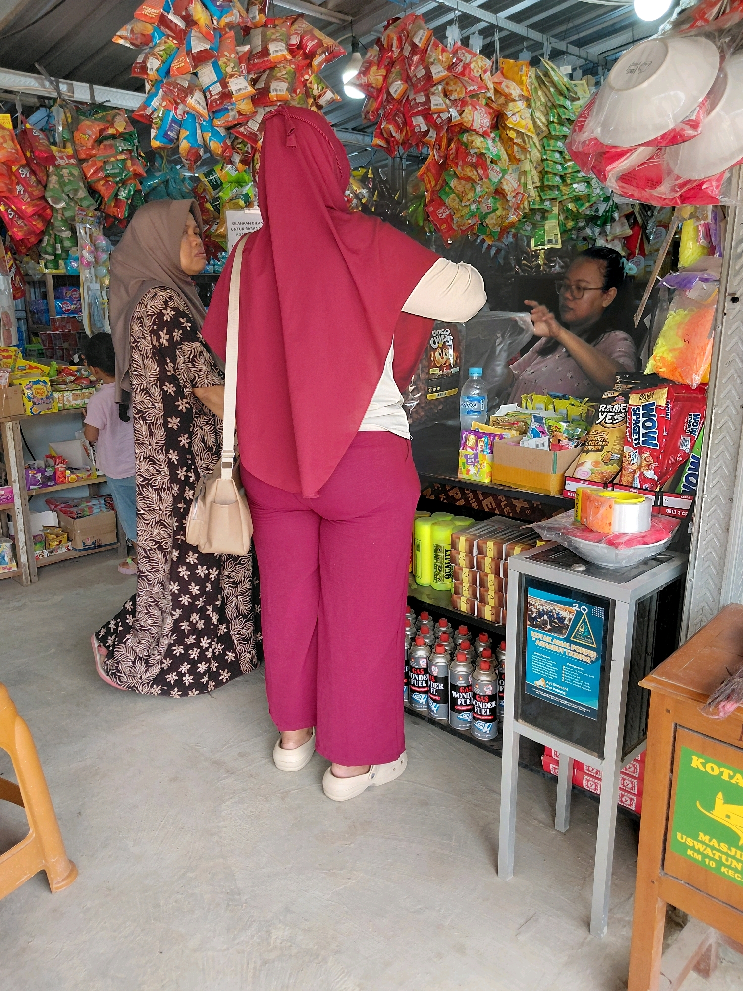 In the city of Persiapan Mulya Raya, Indonesia, a small shop displays a variety of goods. Two women, one wearing a maroon hijab and the other a brown hijab and patterned dress, are visible. The woman in the maroon hijab stands facing away from the camera, while the woman in the brown hijab is turned to her left, looking towards a third person behind the counter. This third person, a woman wearing glasses and a light-colored shirt, is seen from the chest up, appearing to interact with the customers. The shop is filled with shelves stocked with packaged food items, including chips and snacks. Bottles of water and other beverages are also present. A stand with several cans of gas fuel is visible on a lower shelf. The shop appears to be a small, local establishment, possibly a convenience store or warung.