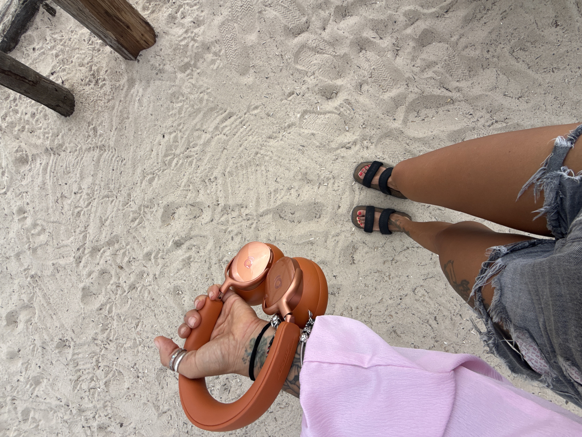 A first-person perspective shot from above shows a person holding orange-colored headphones in their right hand while standing on a sandy beach. The person is wearing a light pink top and denim shorts. Their feet are clad in black sandals. The sand is mostly smooth with some footprints and debris scattered across it. Two wooden posts are visible in the upper right corner of the image, suggesting a structure or barrier on the beach. The overall scene suggests a casual outing on a beach in Choeng Thale, Thailand.