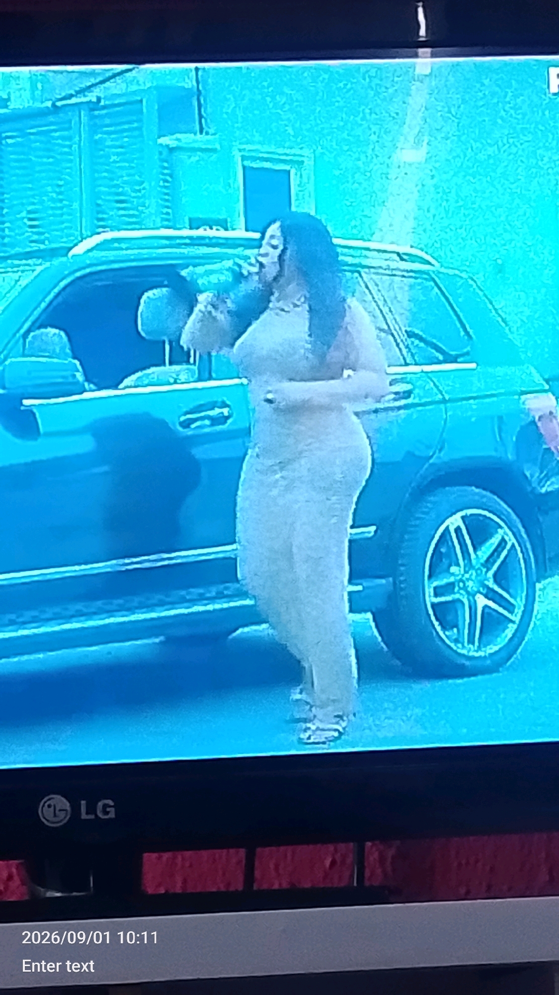 The image shows a woman standing next to a dark-colored SUV. She is wearing a light-colored dress and has dark hair. The woman appears to be holding a bottle to her mouth, possibly drinking. The SUV has tinted windows and chrome rims. In the background, there is a building with multiple windows. The scene is lit by artificial light, casting shadows on the ground. The bottom of the image shows a date and time stamp, "2026/09/01 10:11", and a prompt to "Enter text". The LG logo is visible in the bottom left corner. The image quality is poor, with a grainy, blue-tinted appearance, suggesting it may be a screenshot from a low-resolution security camera or video feed. There is no indication of the specific location in Lokoja, Nigeria.